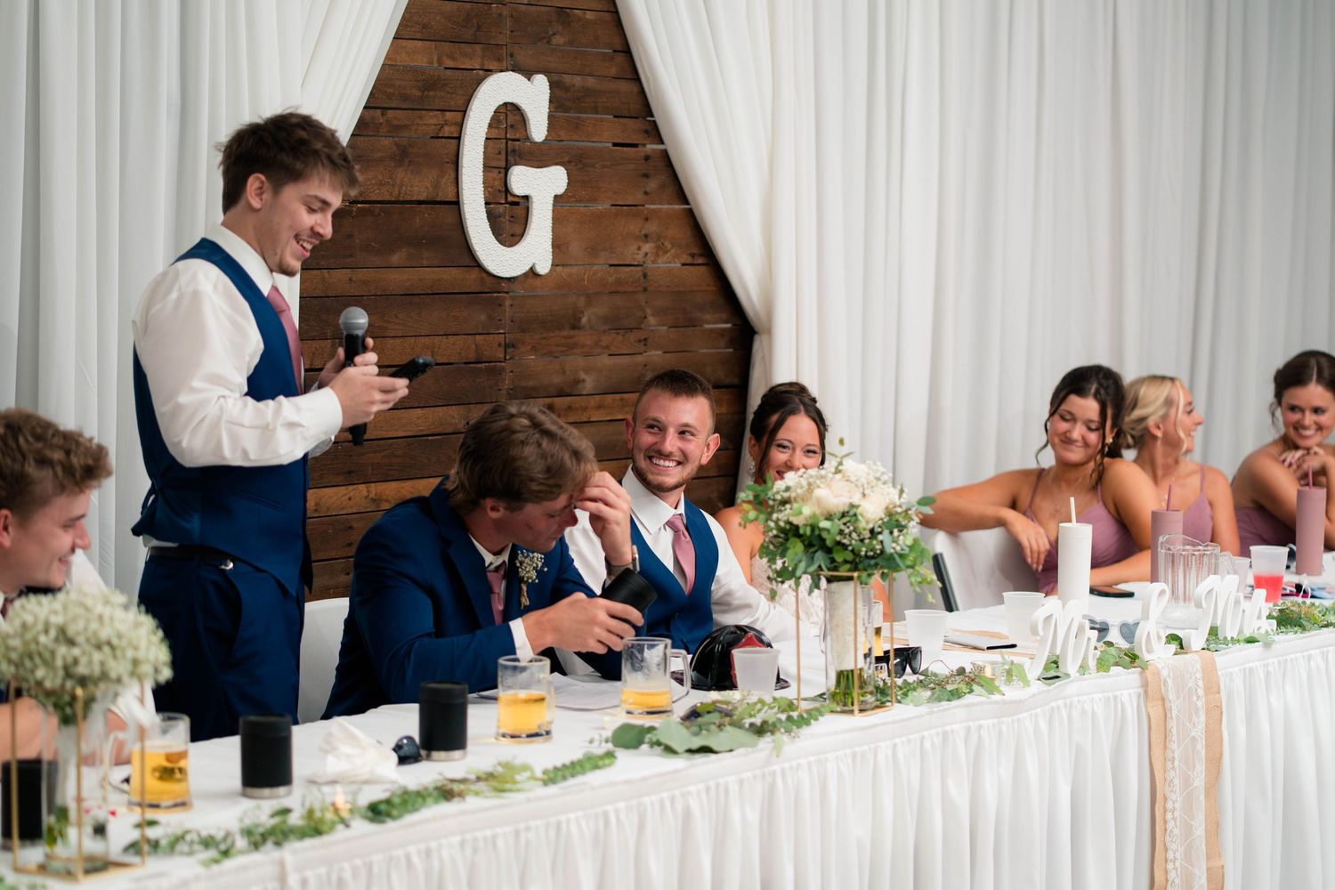 Best man delivers speech at wedding head table while groom laughs with bridal party seated nearby.