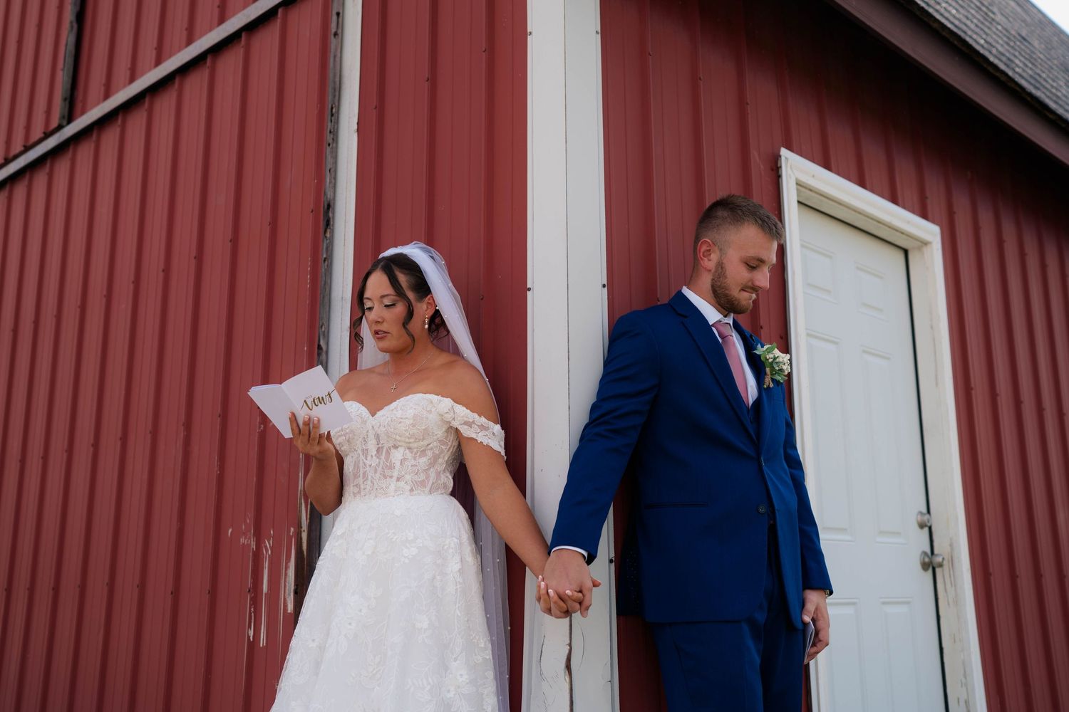 Bride and groom hold hands outside red barn, bride reading vows in off-shoulder lace gown and veil.