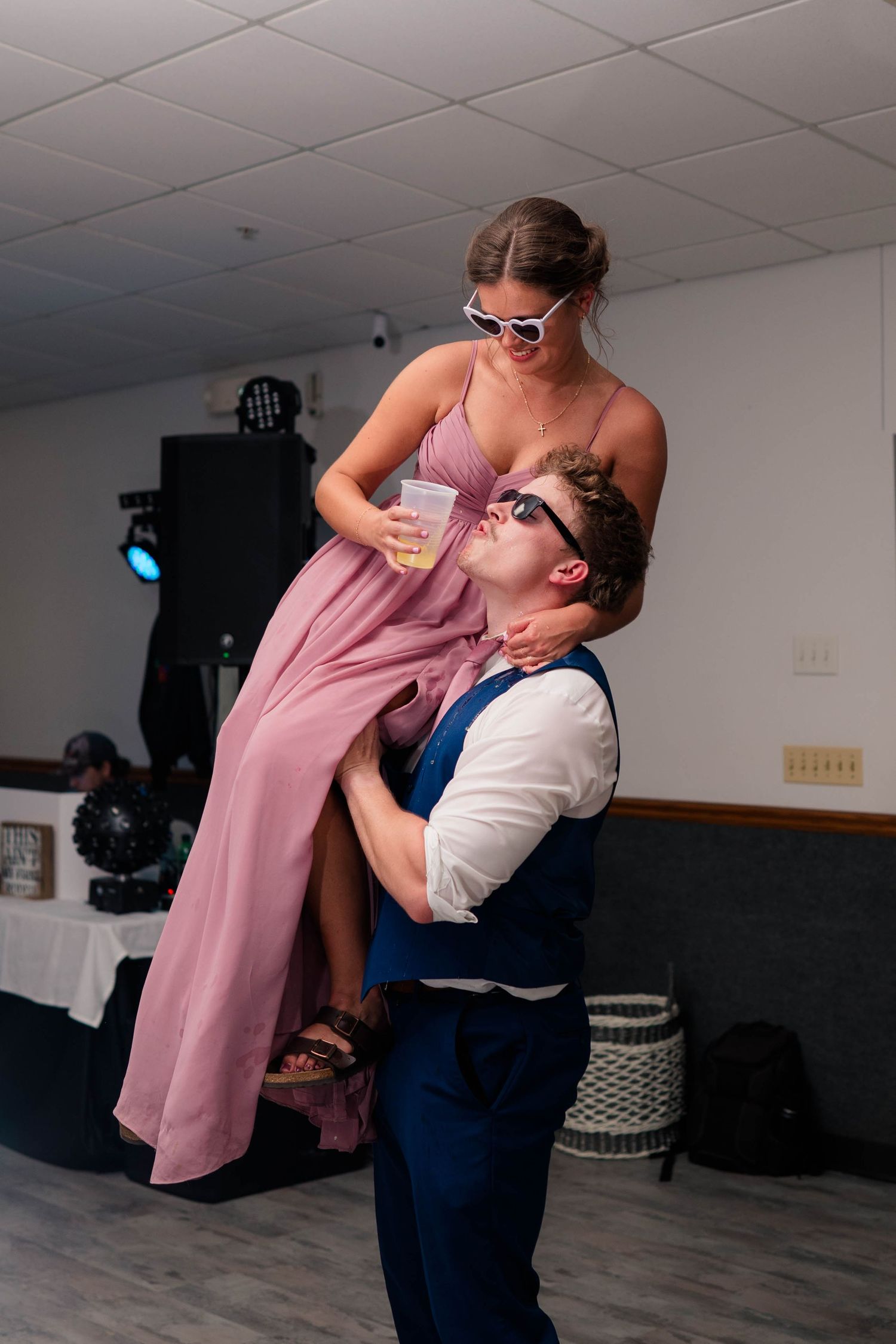 A man in a blue suit lifts a woman in a pink gown at a wedding reception while both wear fun sunglasses.