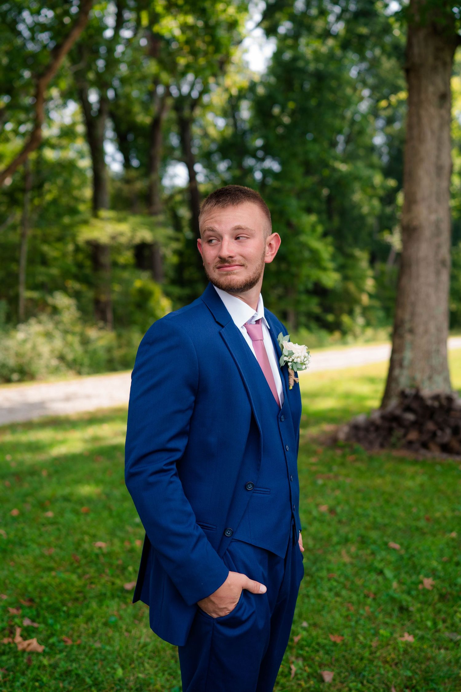 Dapper groom in navy blue three-piece suit with pink tie and boutonniere poses outdoors in green park.