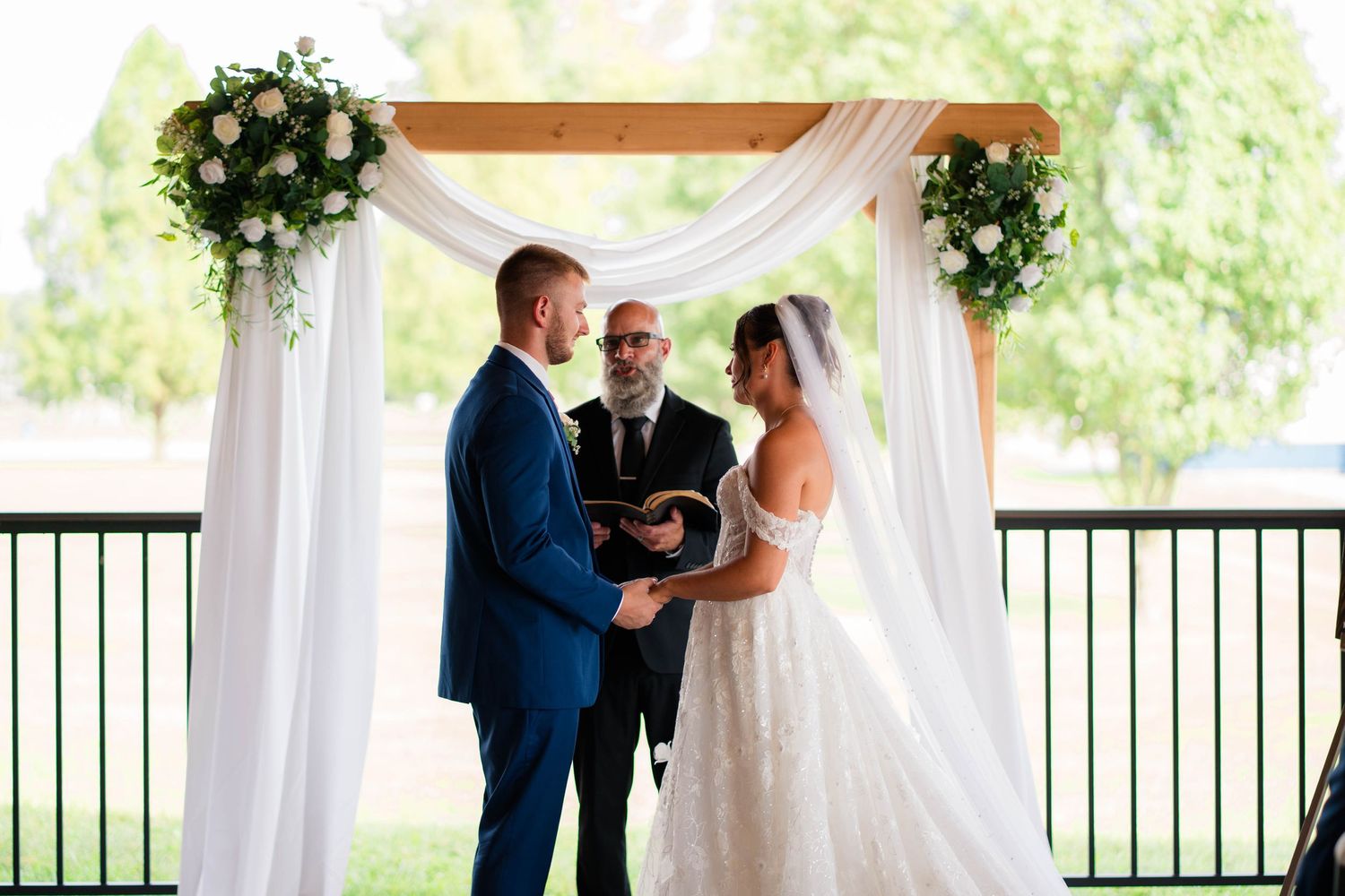 Bride and groom exchanging vows under floral wooden arch with officiant at wedding.