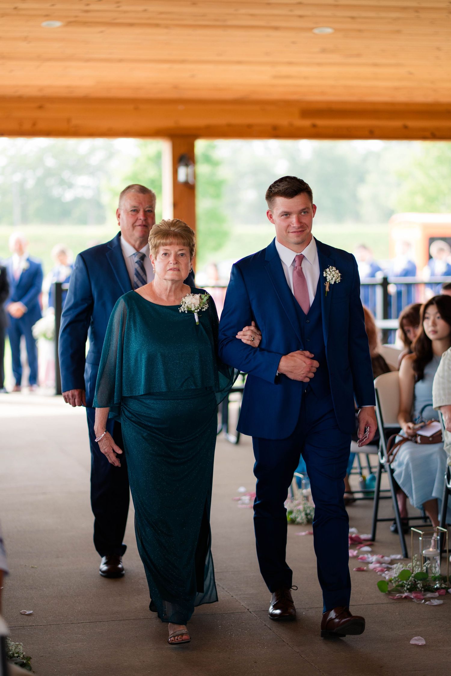 Groom in navy suit escorts older woman in teal dress down wedding ceremony aisle under wooden pavilion.