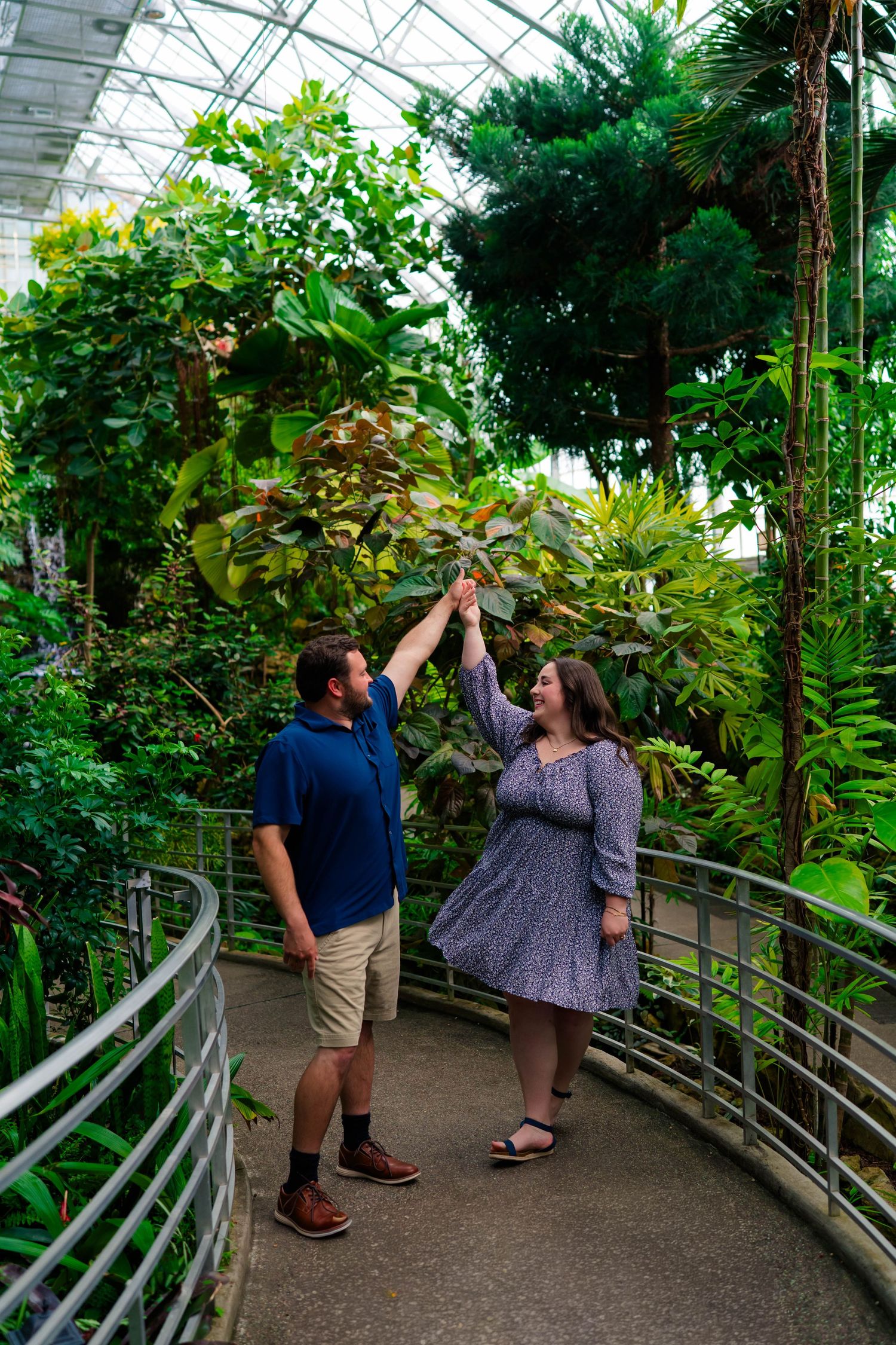 Couple playfully reaching up to touch tropical foliage on a walkway in a greenhouse.