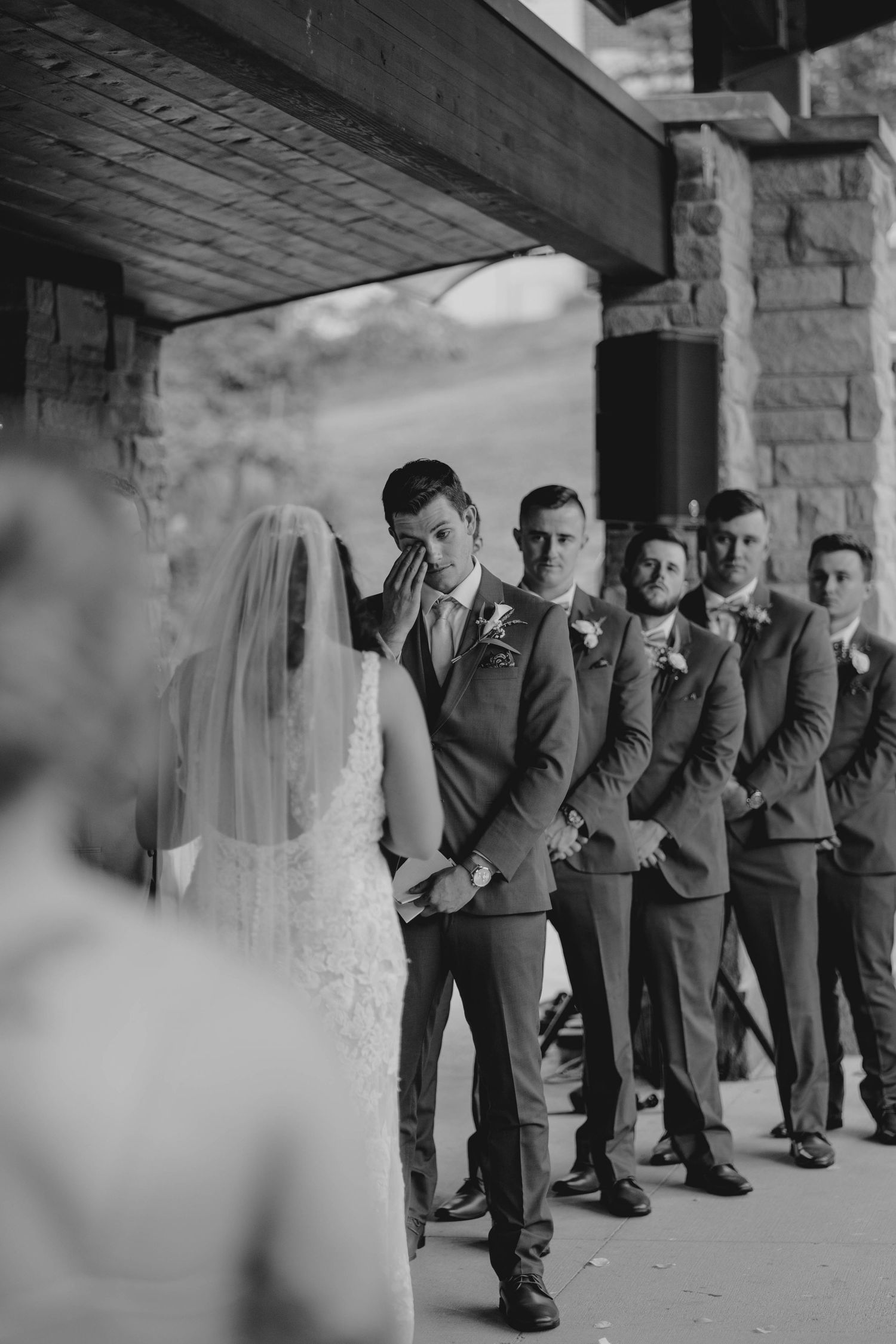 Emotional groom wipes tears as bride walks down the aisle in black and white photo.
