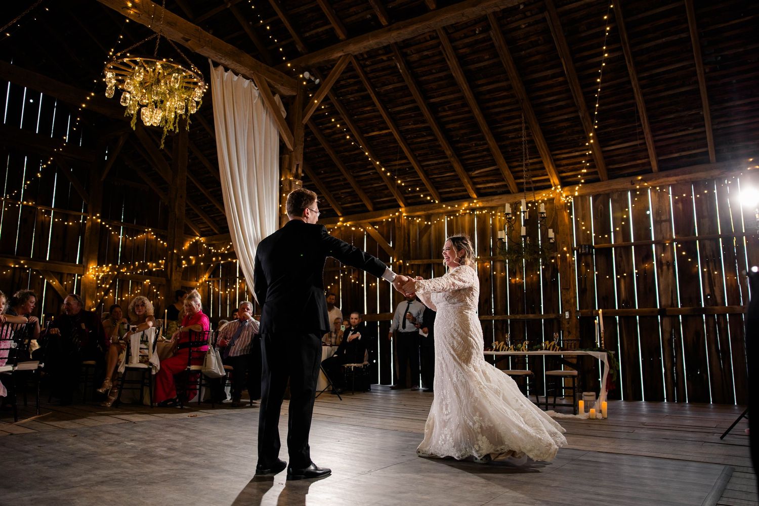 Bride and groom share elegant first dance in rustic barn decorated with twinkling string lights.