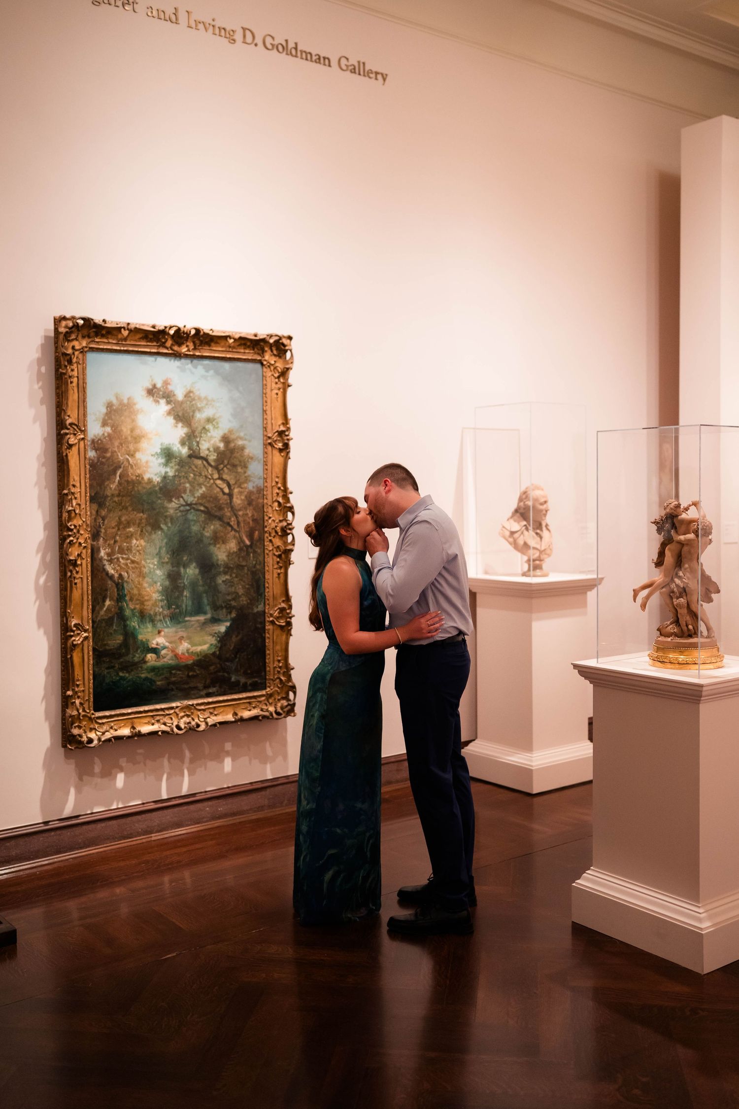 Couple sharing a romantic kiss in a Cincinnati Art Museum gallery with a gold-framed landscape painting and sculptures.