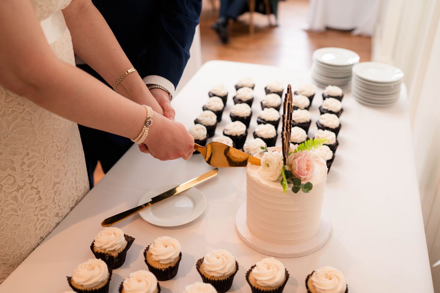Bride and groom cutting white wedding cake surrounded by rows of frosted chocolate cupcakes at reception.
