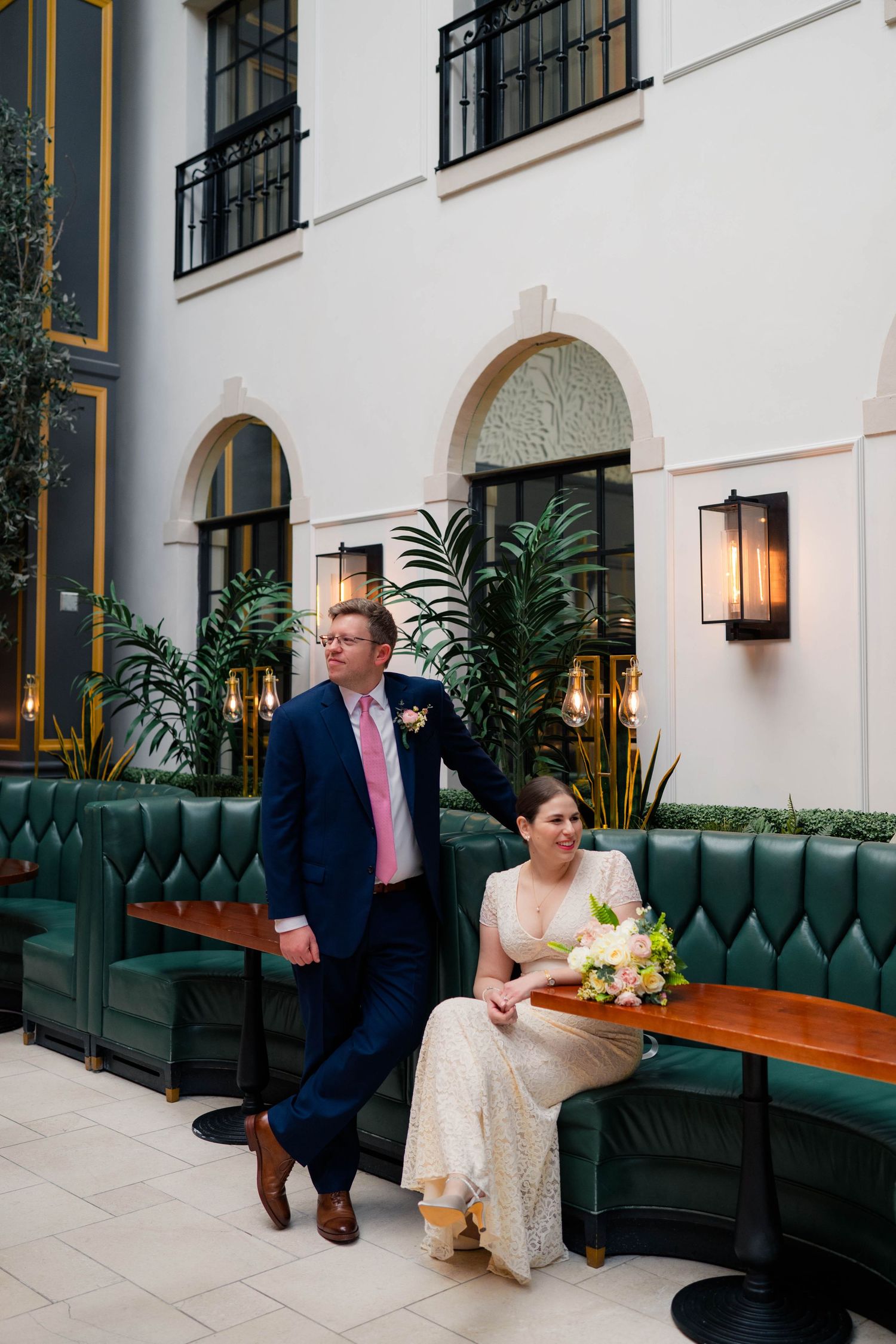 Wedding couple poses in stylish green velvet booth at upscale venue, bride holding bouquet.