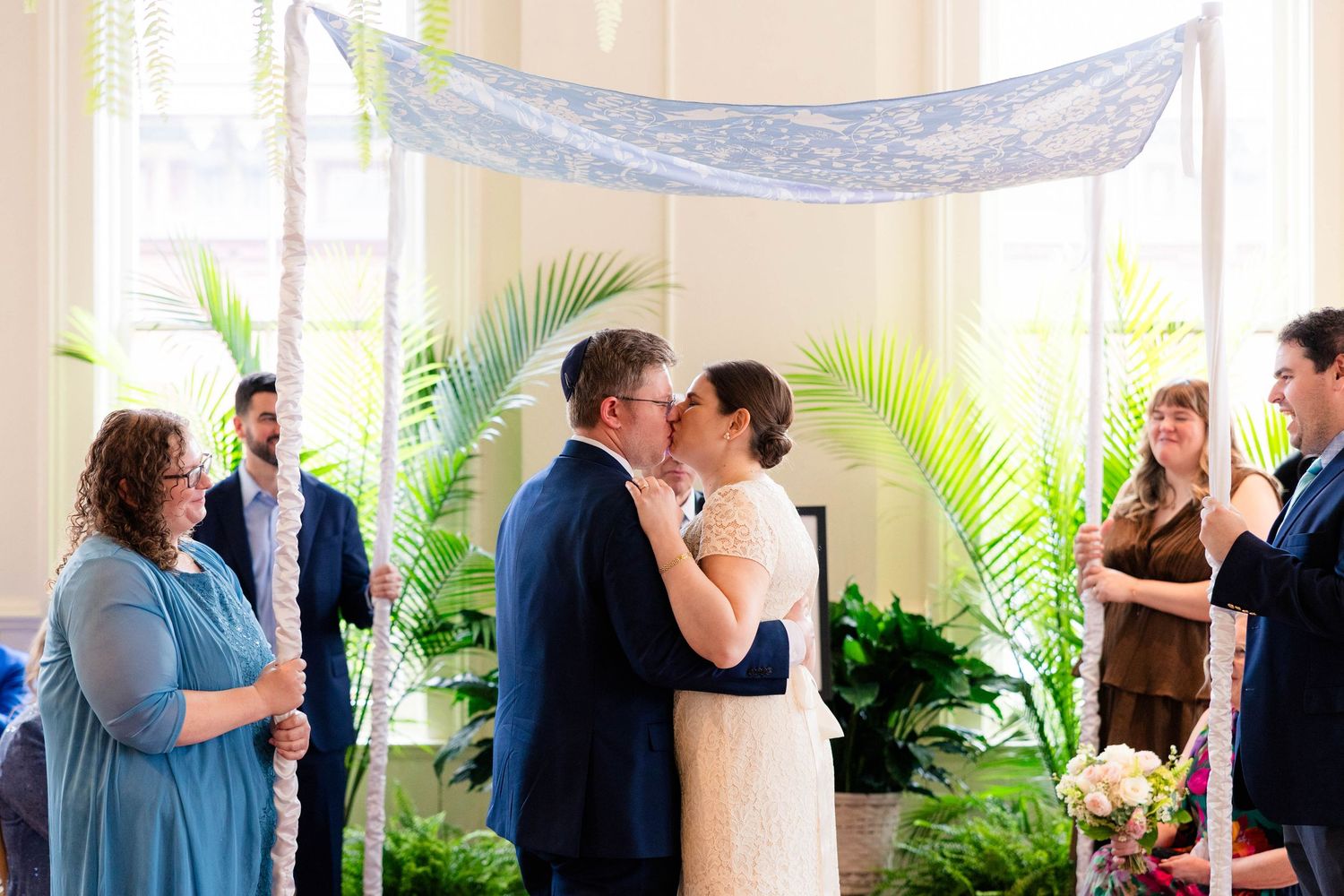 Bride and groom share first kiss at wedding altar decorated with palm leaves and draped fabric.