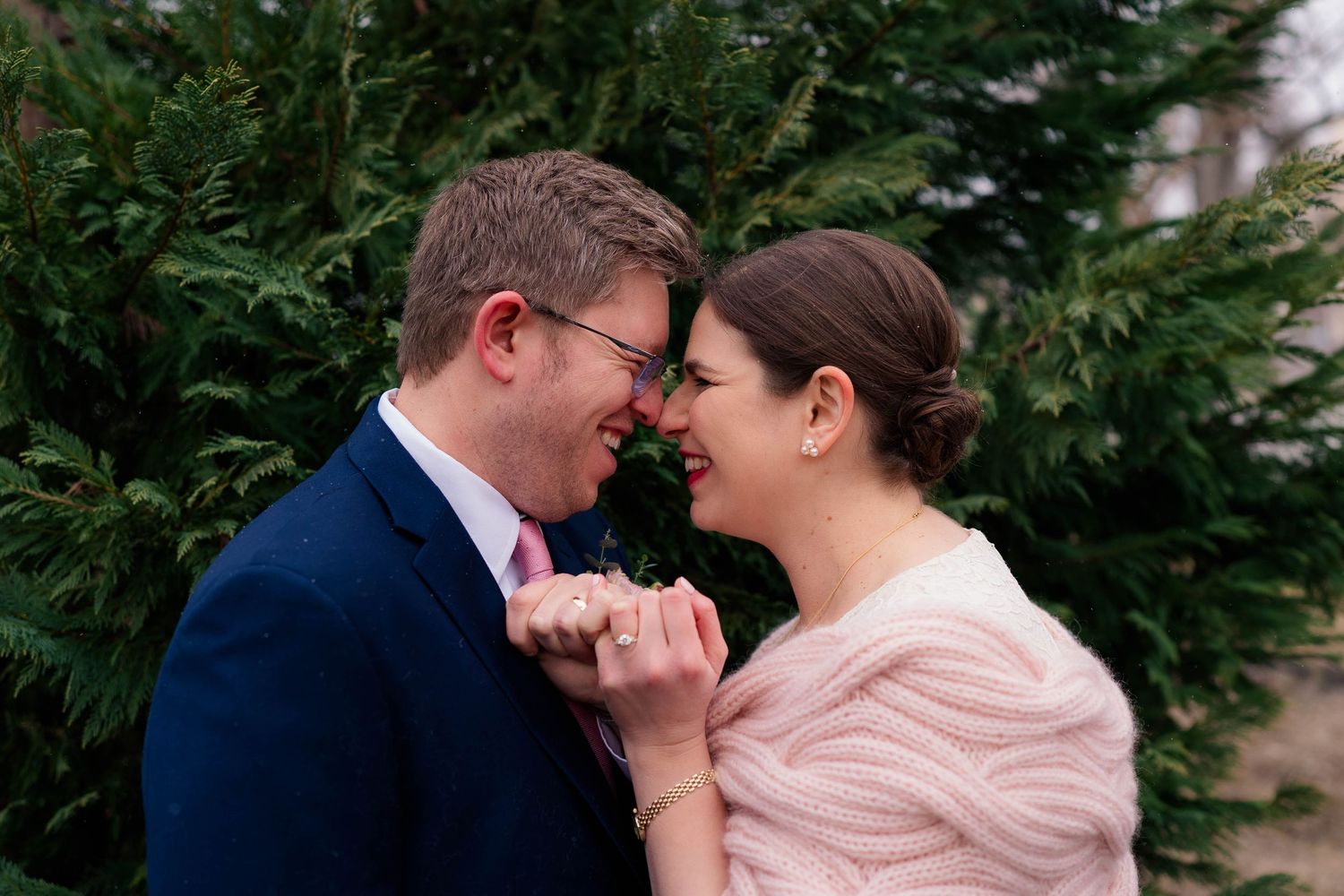 Bride and groom touch foreheads and smile while holding hands in front of lush evergreen trees.