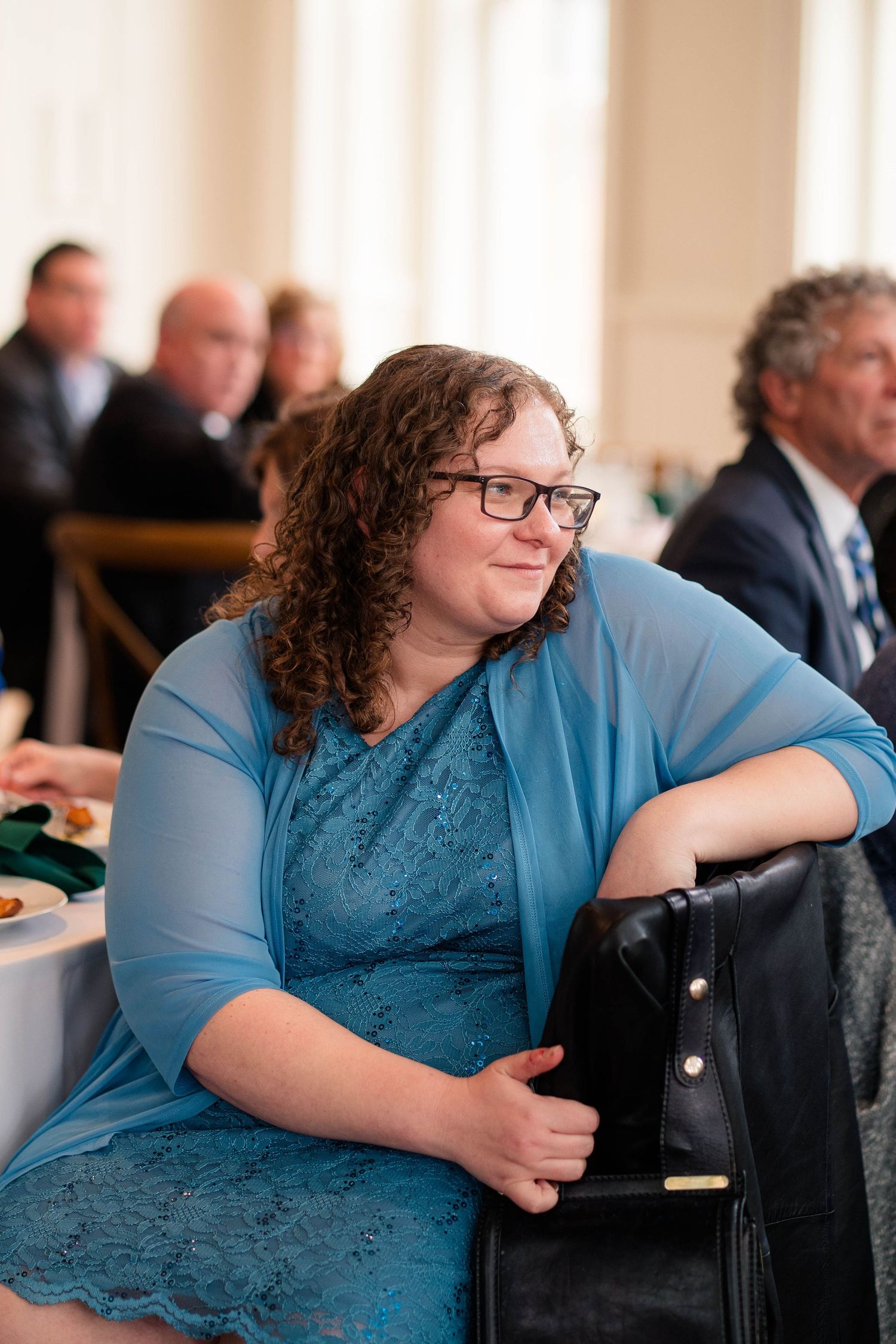 Woman in blue lace dress with glasses smiling at wedding reception while holding black purse.