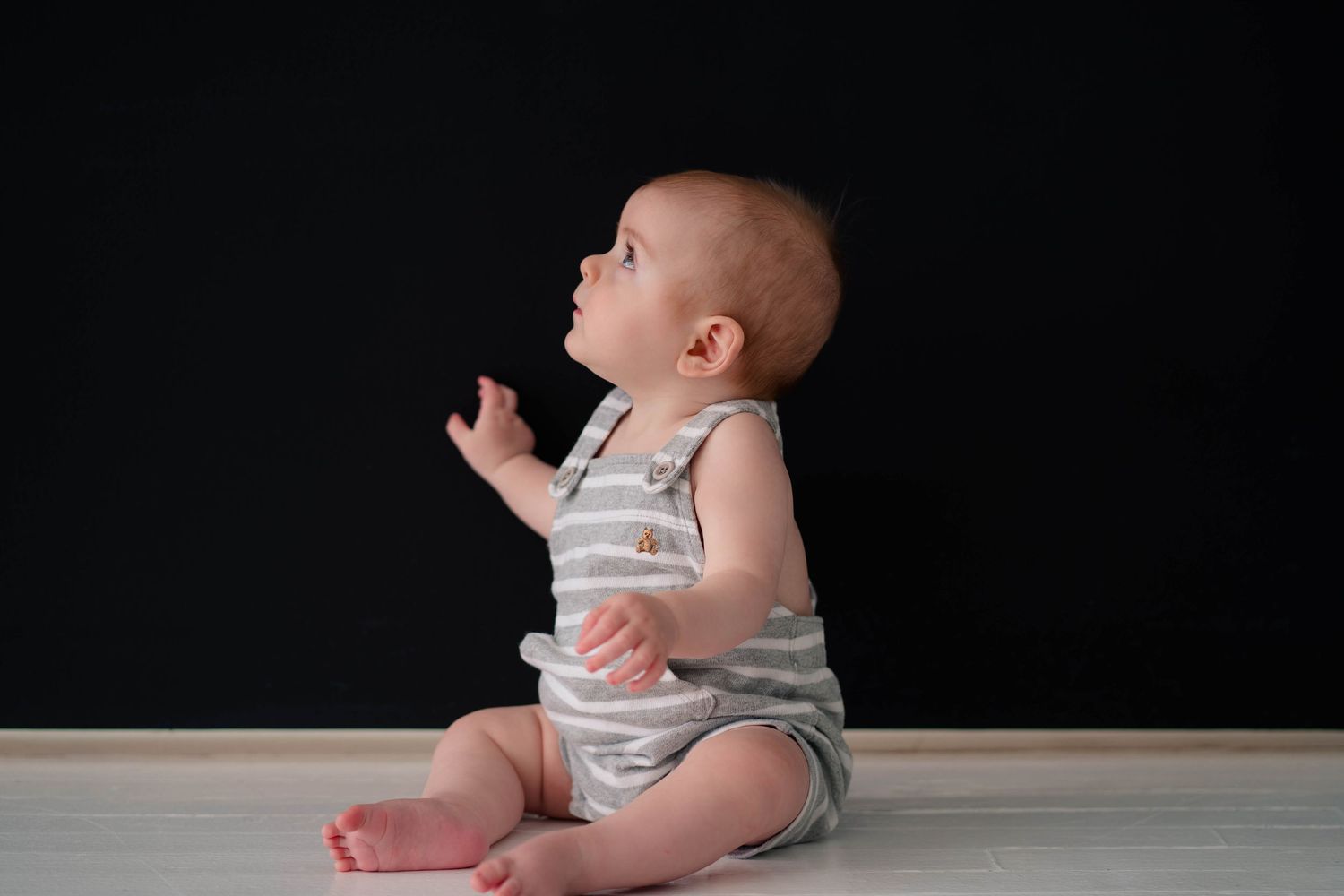 An adorable baby in a striped romper sits and reaches up against a dramatic black background.