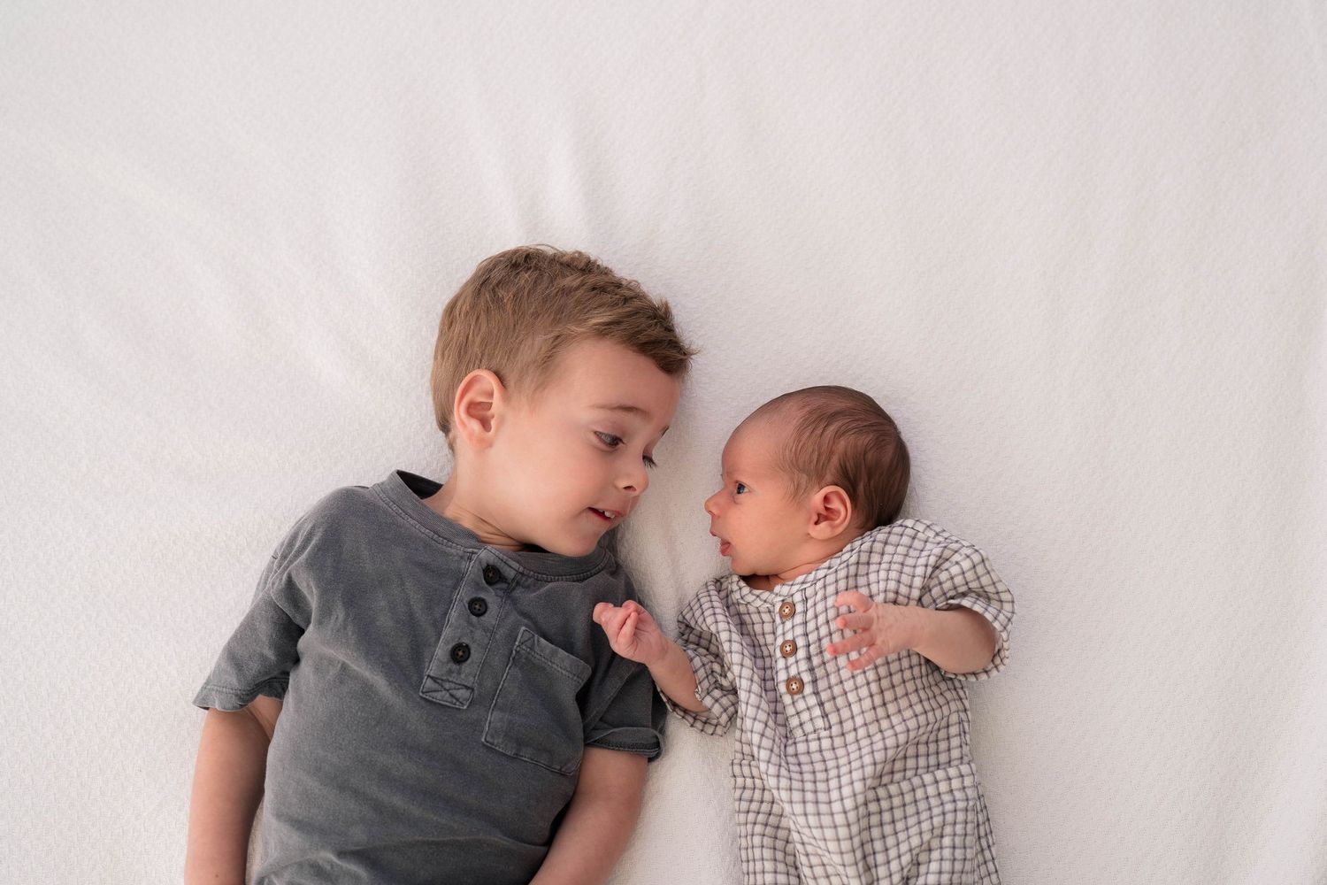 Young boy in gray shirt lying next to newborn baby in checkered onesie on white background.