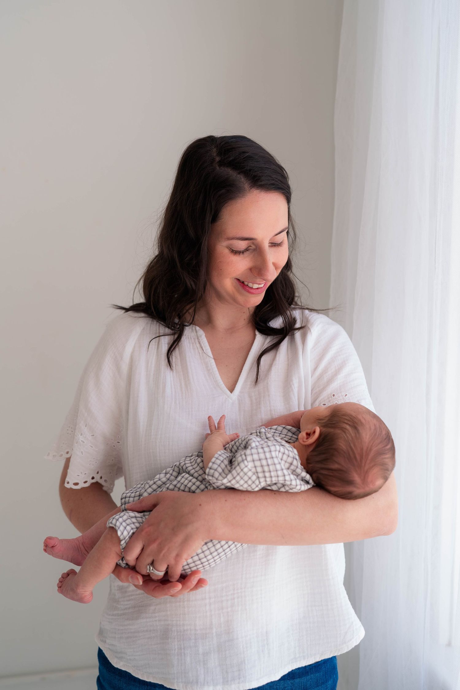 Smiling dark-haired mother in white top cradling newborn baby in bright airy room.