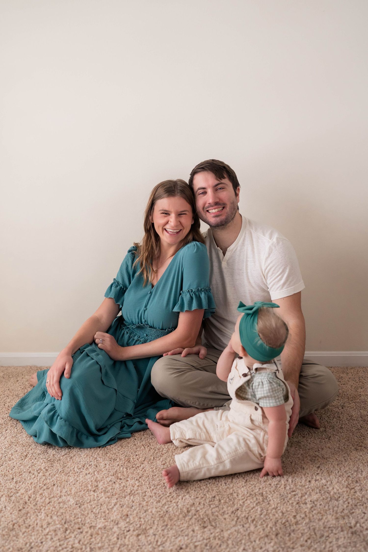 Happy couple sitting on carpet floor with their baby in a teal dress and white shirt studio portrait.