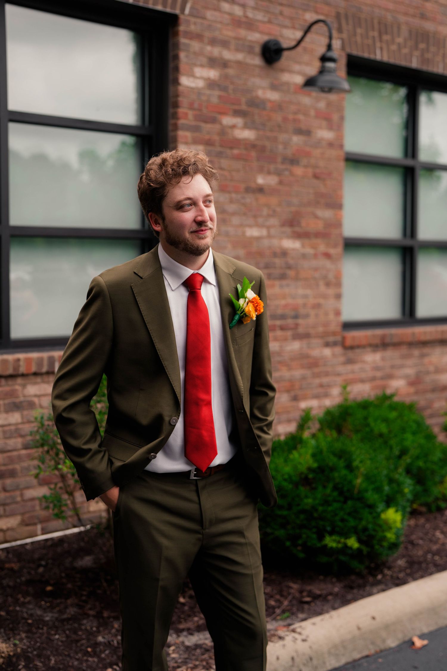 Groom in olive green suit and red tie with floral boutonniere stands outside brick building.