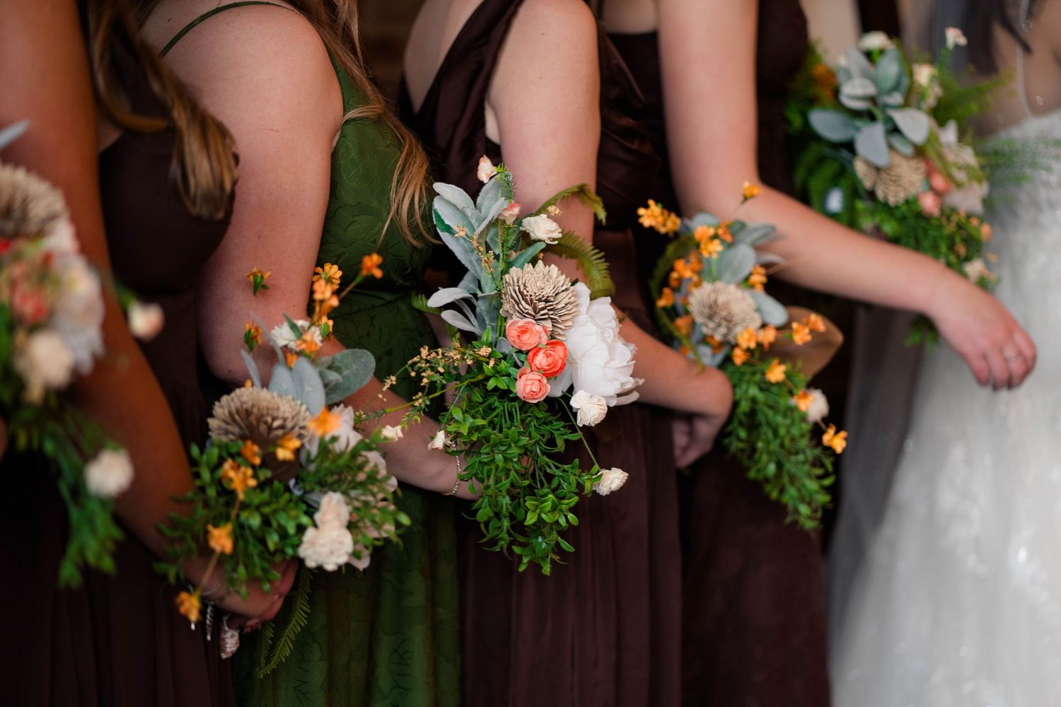 Bridesmaids in brown dresses holding rustic bouquets with peach roses, orange flowers, and greenery at a wedding.