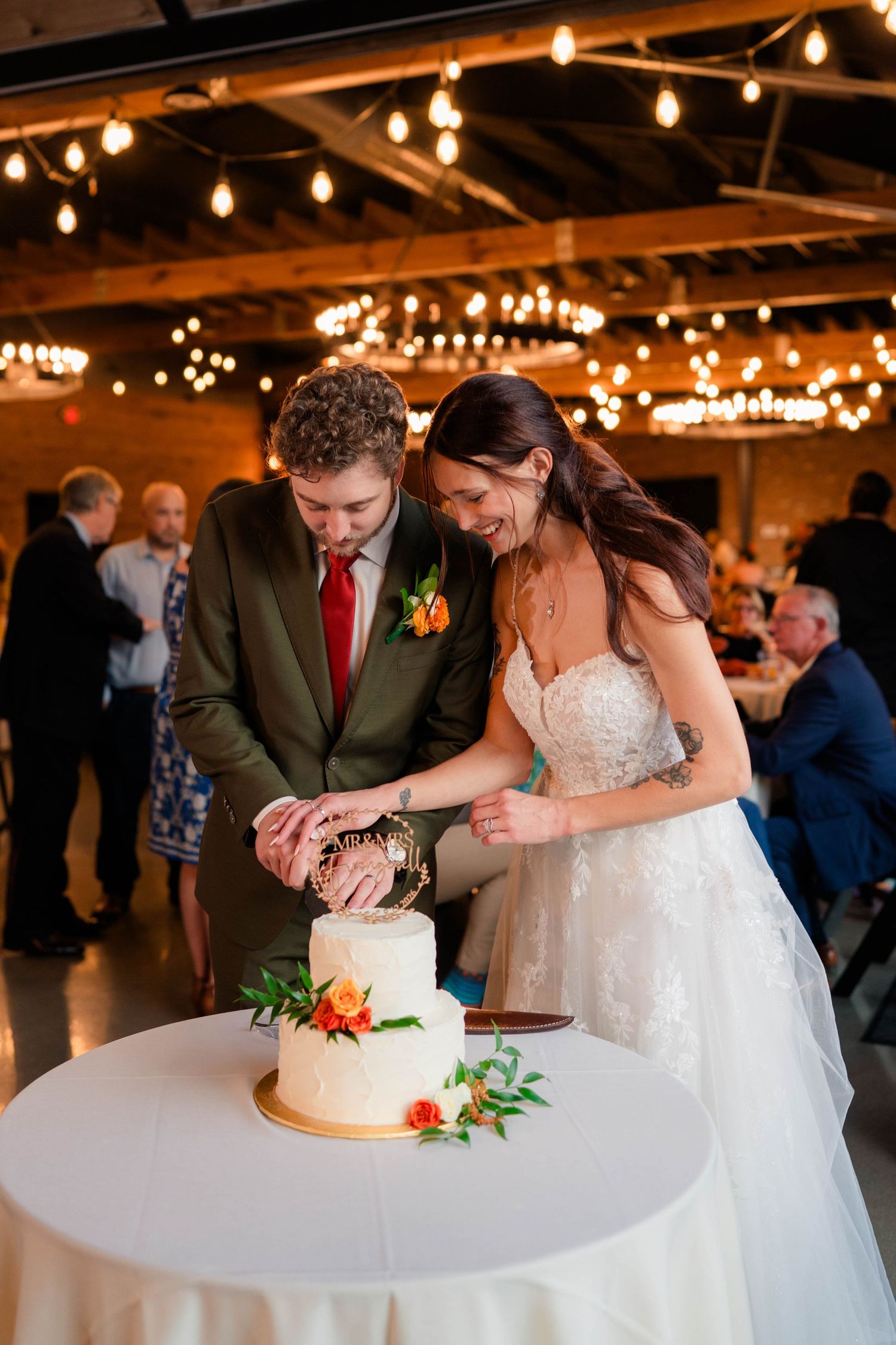 Bride and groom cut two-tier white wedding cake with floral decor under string lights.