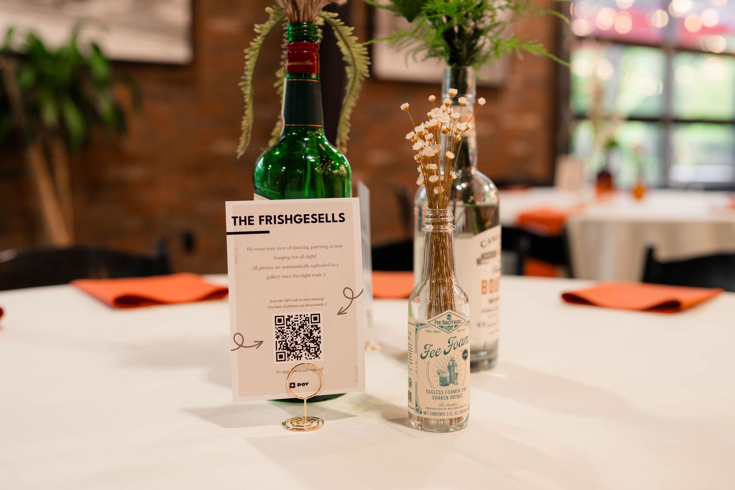 Wedding reception table centerpiece featuring liquor bottles with flowers and Frishgesells QR code sign.