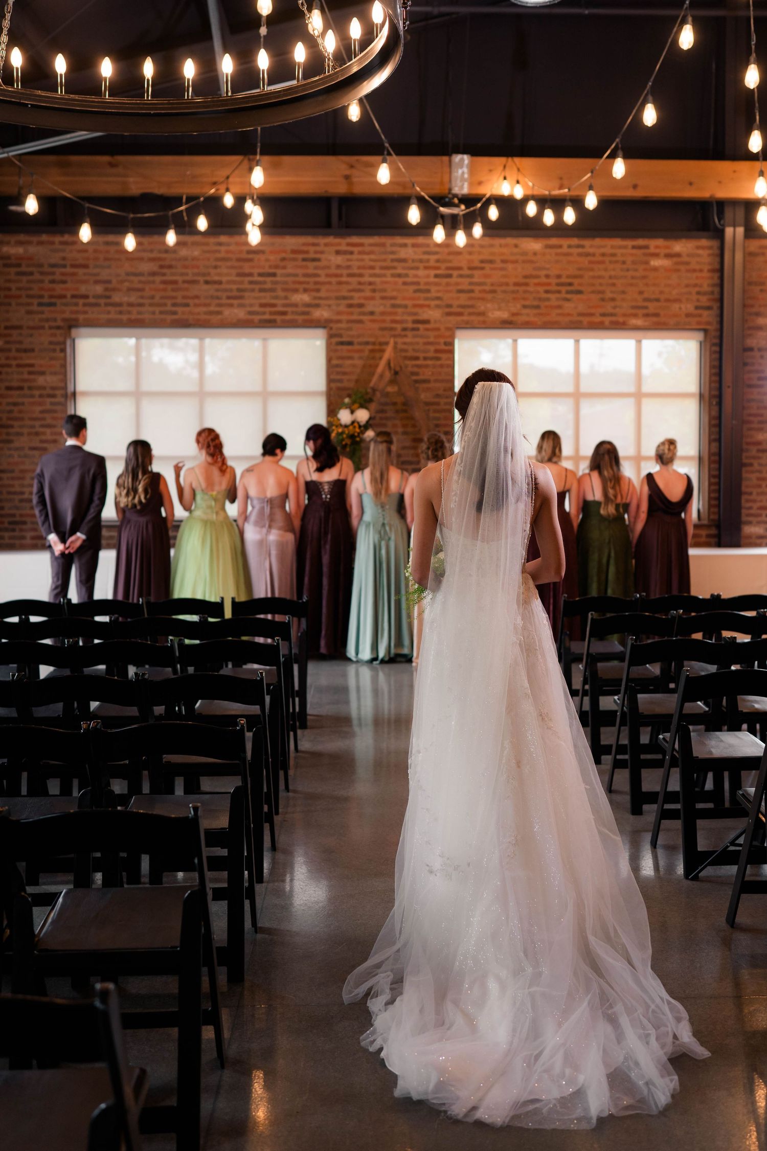 Bride in white gown and veil walking toward wedding party in a rustic brick venue with string lights.