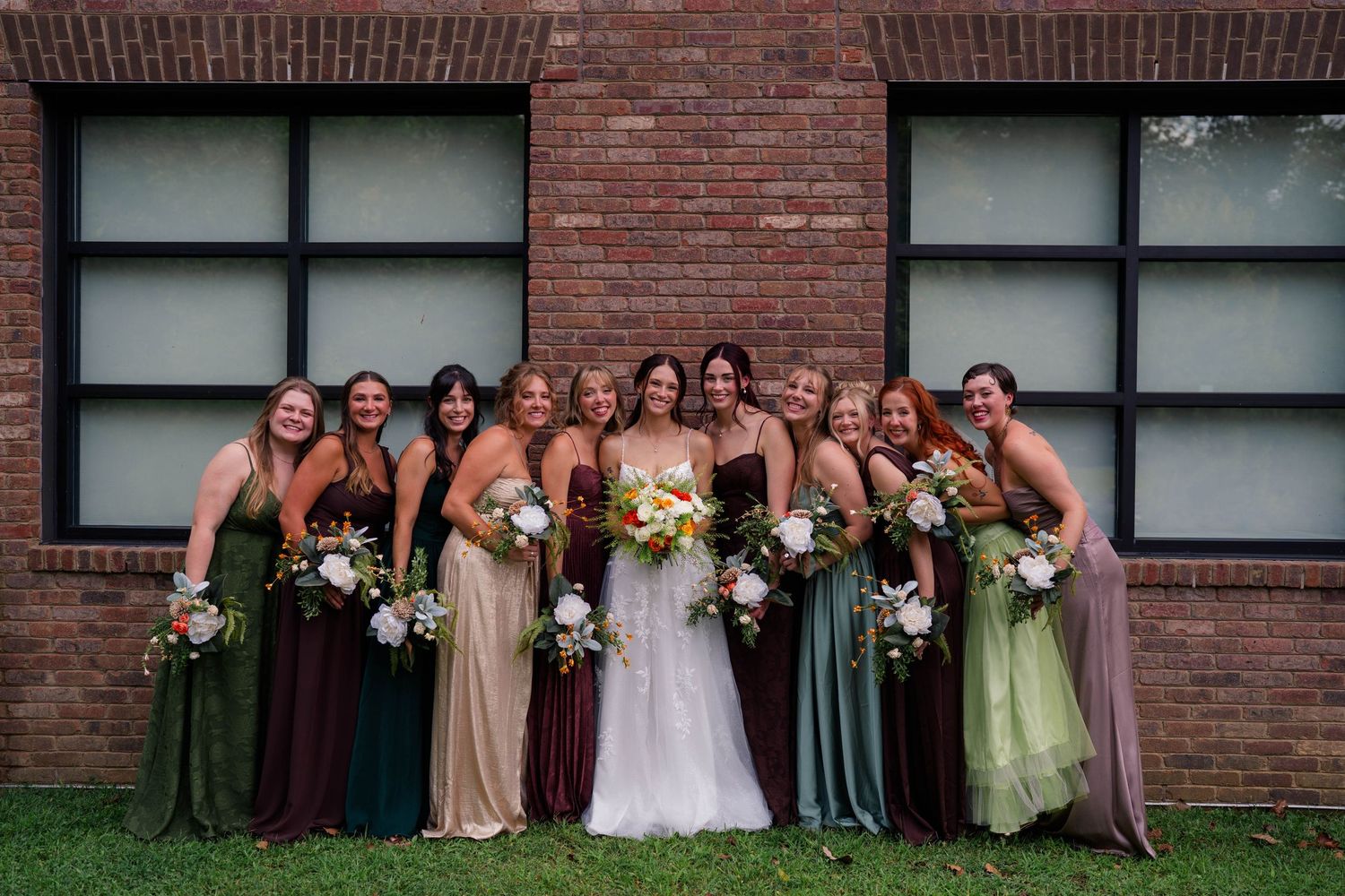 Bride poses with nine bridesmaids in mismatched earth-tone gowns outside rustic brick wedding venue.