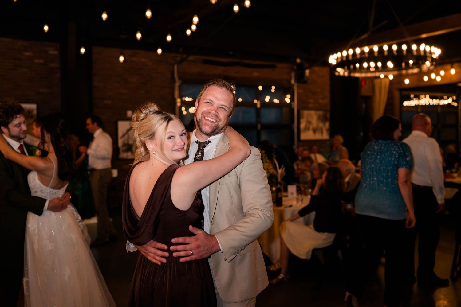 Smiling couple embracing on wedding reception dance floor in warmly lit industrial venue at night.