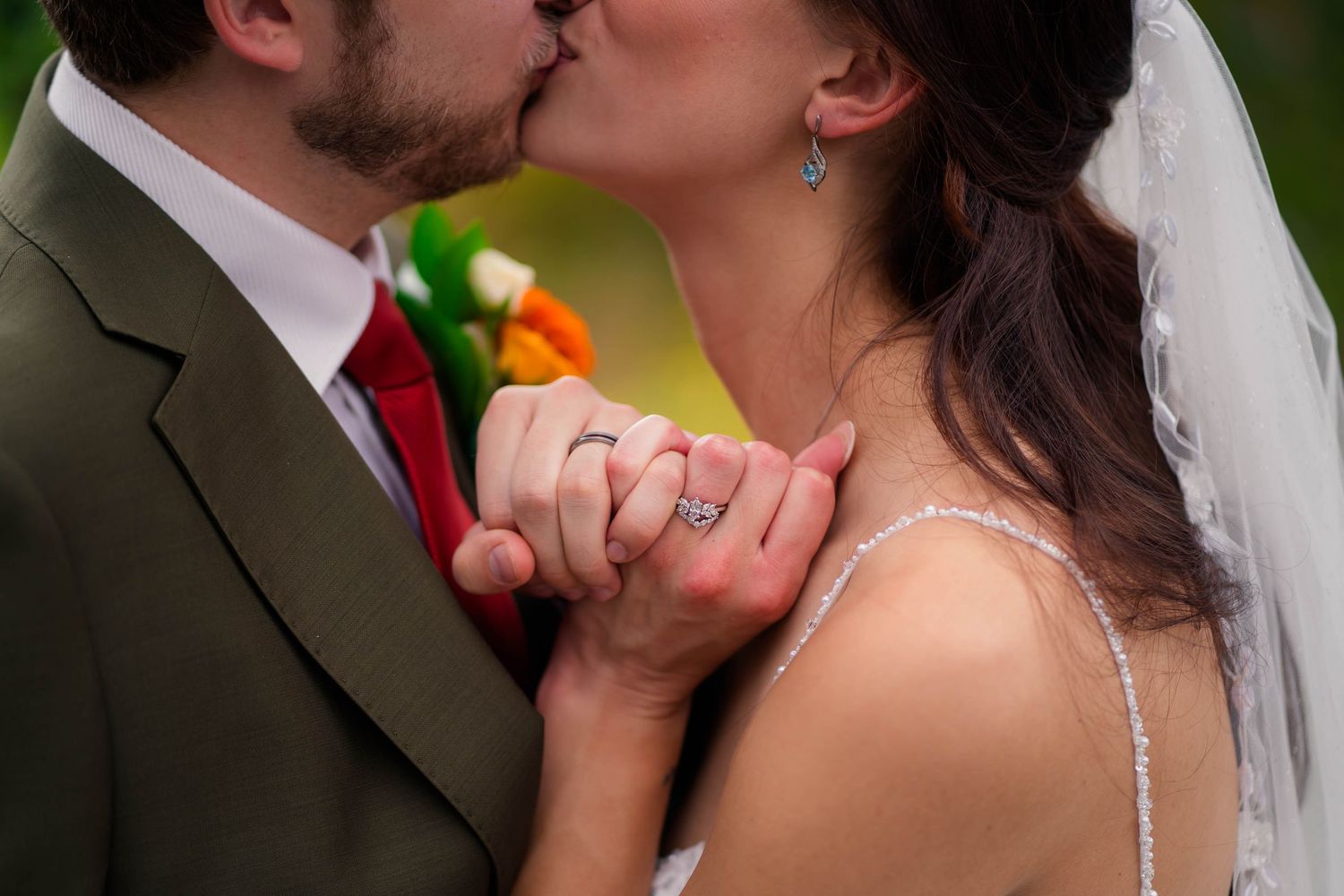 Bride and groom kissing while holding hands showing wedding ring with colorful bouquet.