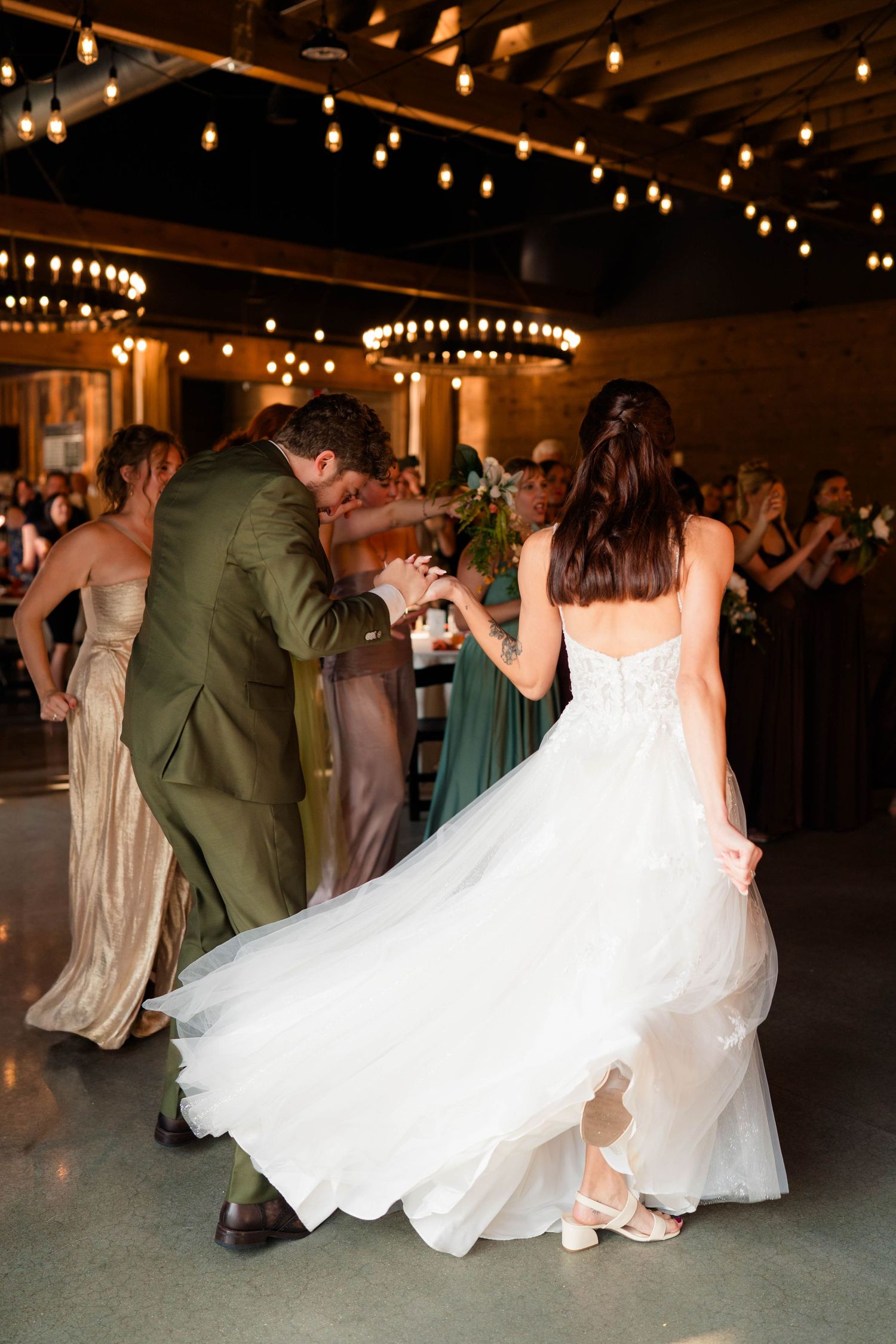 Bride in white gown and groom in olive suit dance surrounded by guests at wedding.