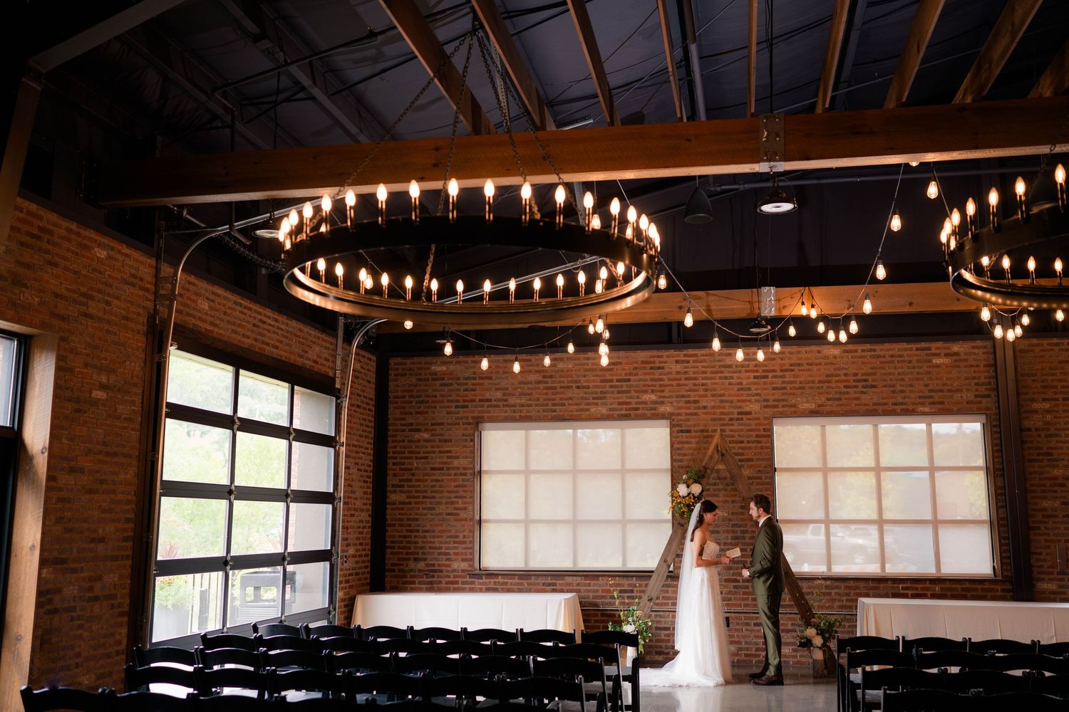 Bride and groom share first kiss under elegant circular Edison bulb chandeliers in industrial brick venue.