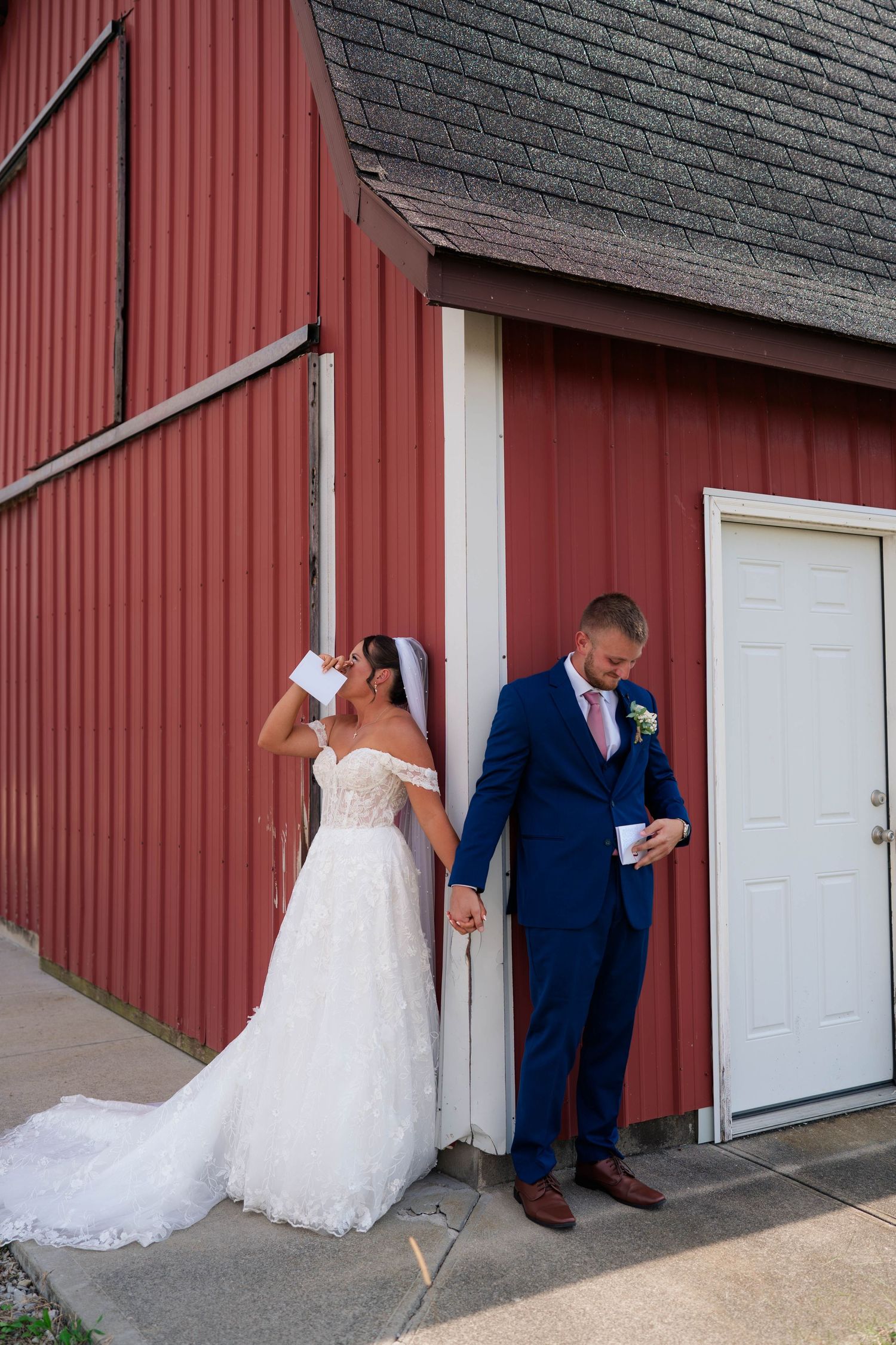 Bride in lace gown and groom in blue suit hold hands reading vows beside a red barn door.