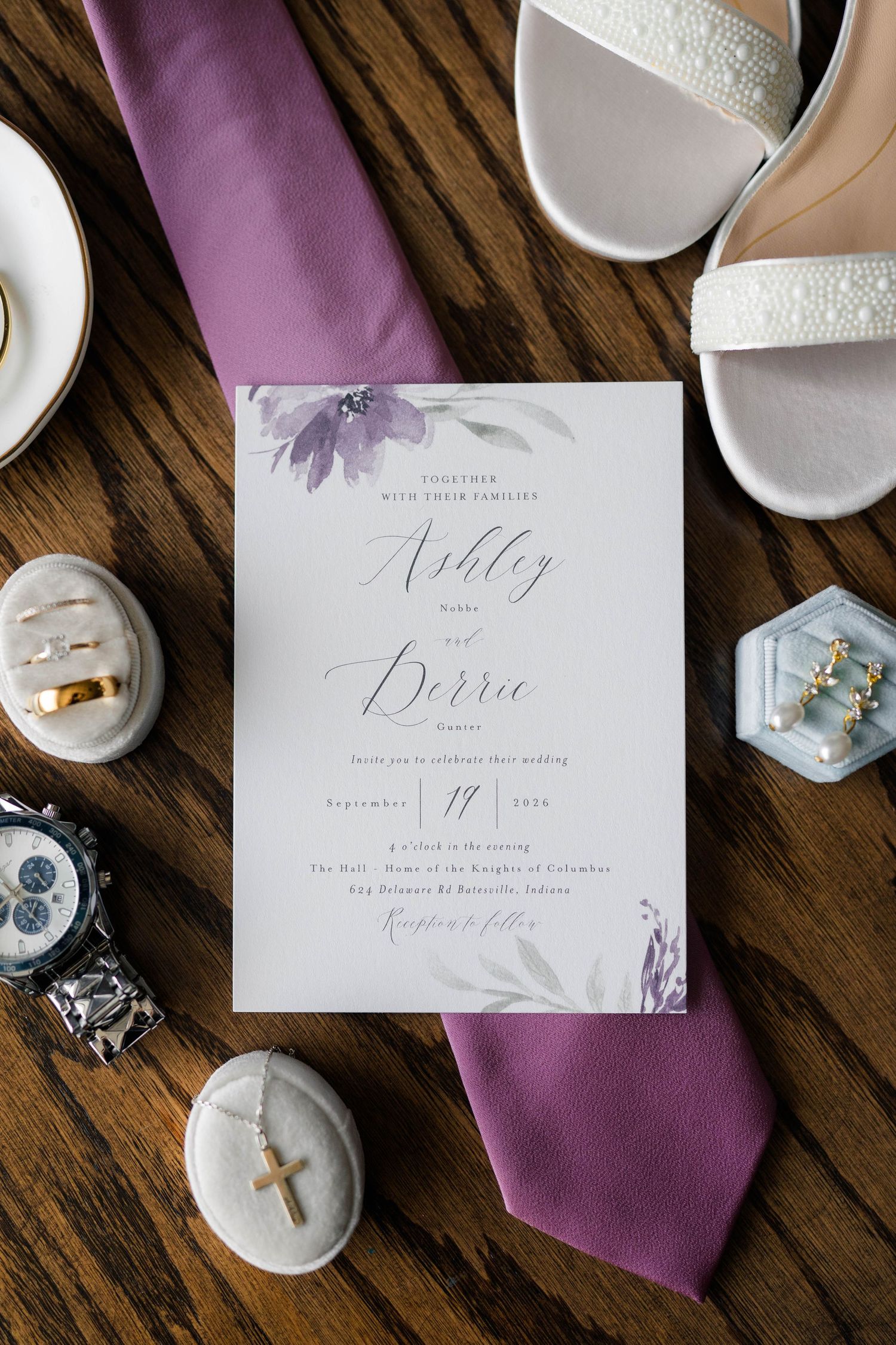 Wedding invitation for Ashley and Derric with purple floral design, surrounded by rings, tie, and bridal accessories.