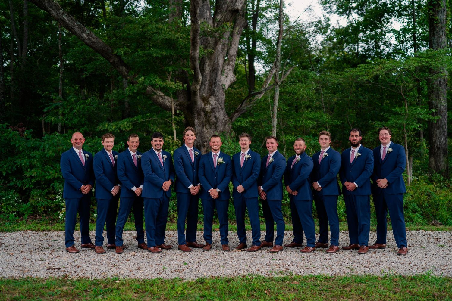 Groom and groomsmen in matching navy blue suits with pink ties pose in a line outdoors at wedding.