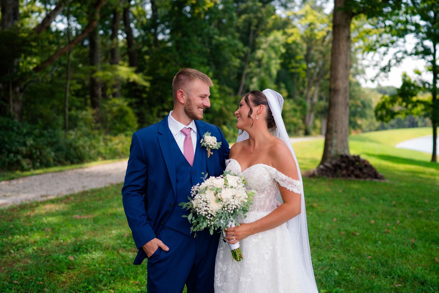Smiling bride and groom in navy suit share a romantic outdoor moment, bride holding white floral bouquet.
