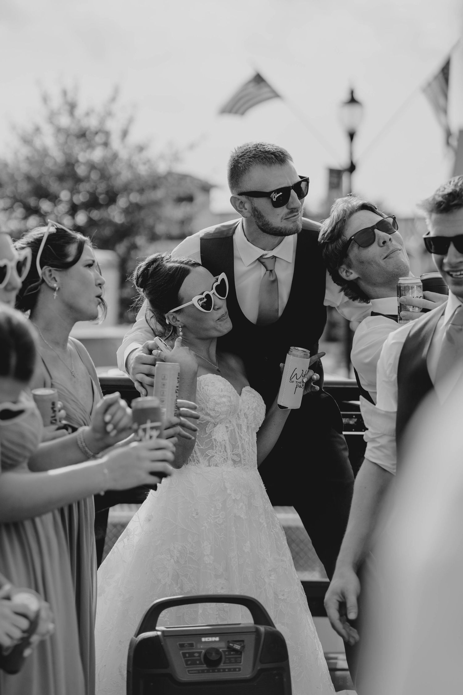 Black and white photo of bride and groom in sunglasses celebrating with wedding party outdoors.