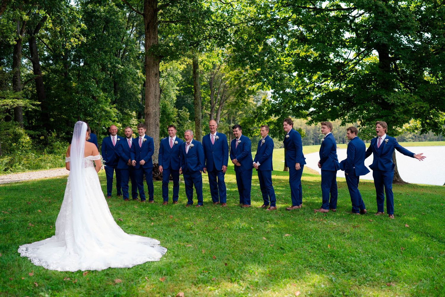 Bride in white gown faces groomsmen in navy suits on a sunny green lawn during wedding party photos.