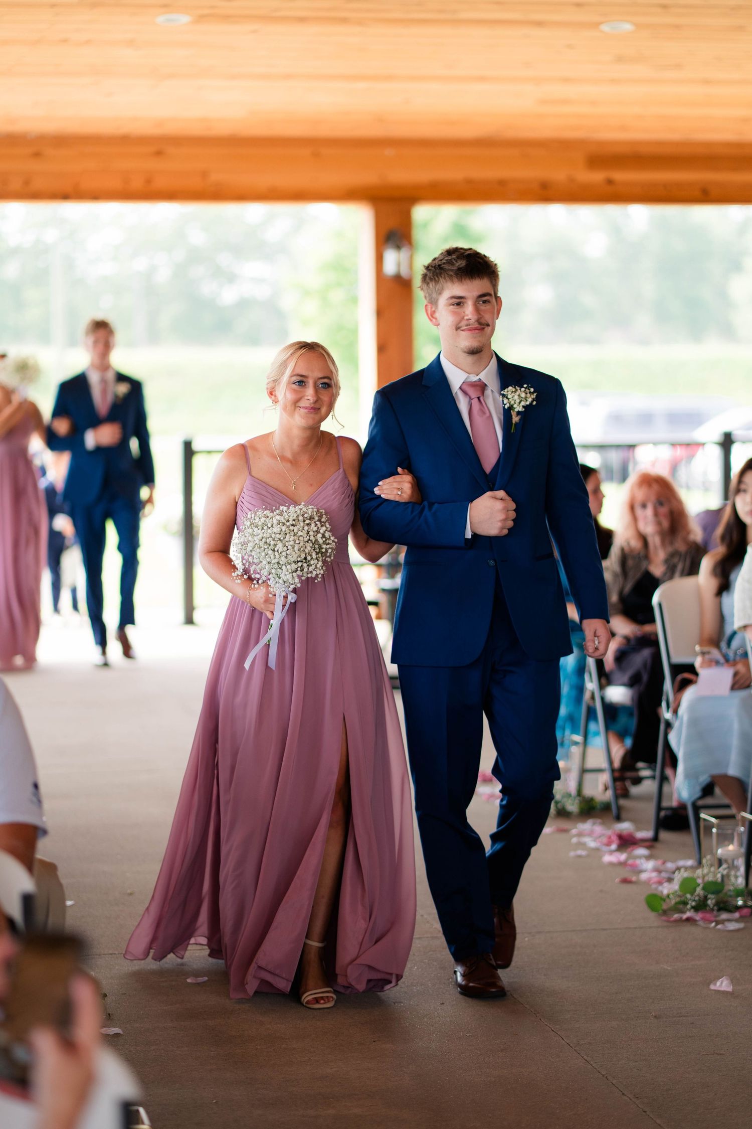 Bridesmaid in mauve gown walks down aisle with groomsman in navy suit at outdoor wedding.