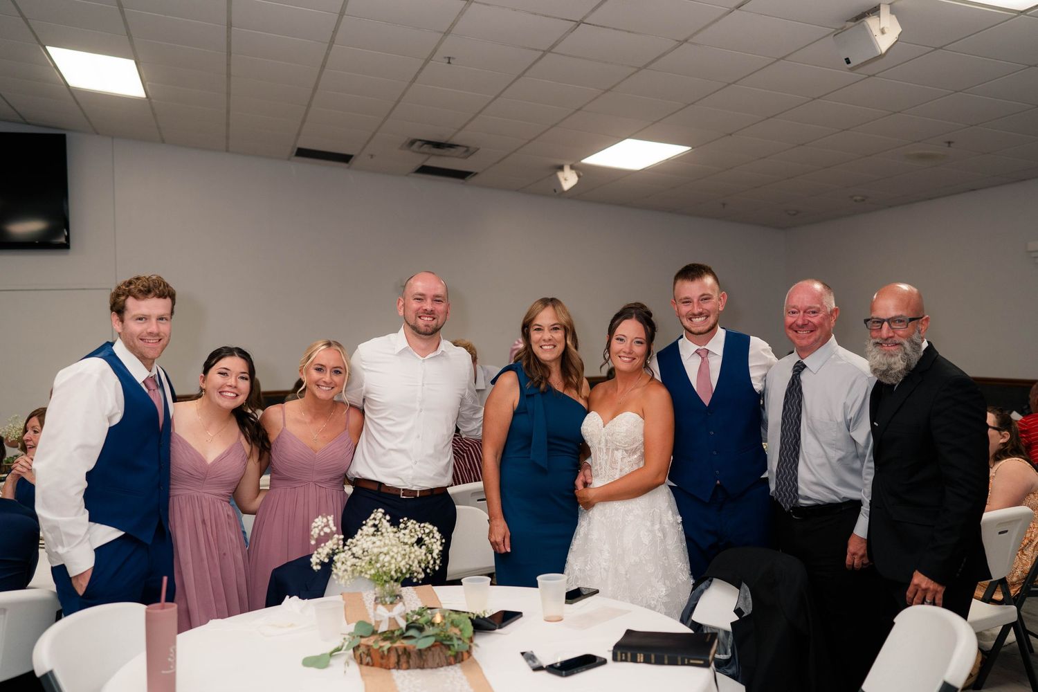 Wedding party and guests pose together smiling at a decorated reception venue with white tablecloths.