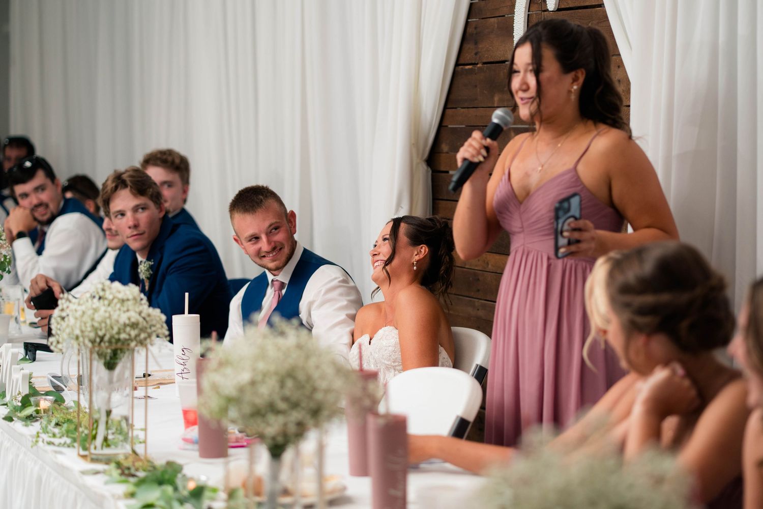 Bridesmaid gives emotional wedding speech as bride and groom smile at elegantly decorated reception table.