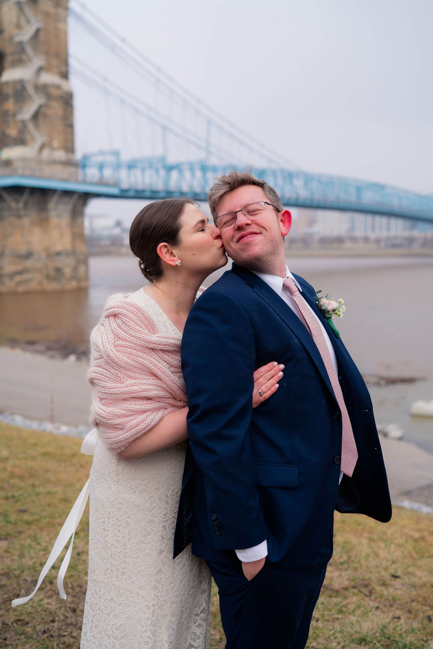 Bride kissing groom on the cheek by a blue bridge, both dressed in wedding attire on a cloudy day.