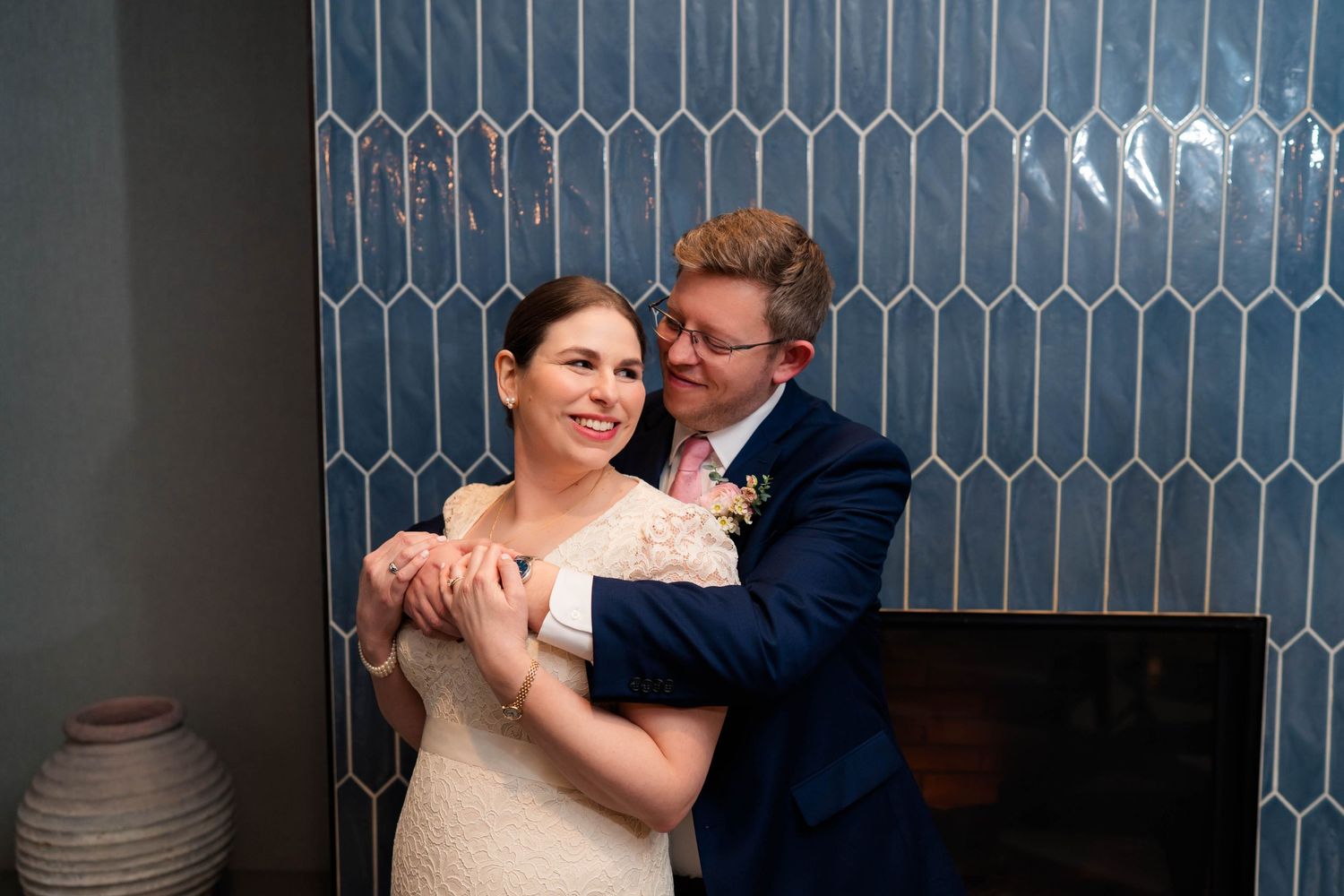 Groom in navy suit embracing bride in lace dress against a blue geometric tile wall at their wedding.