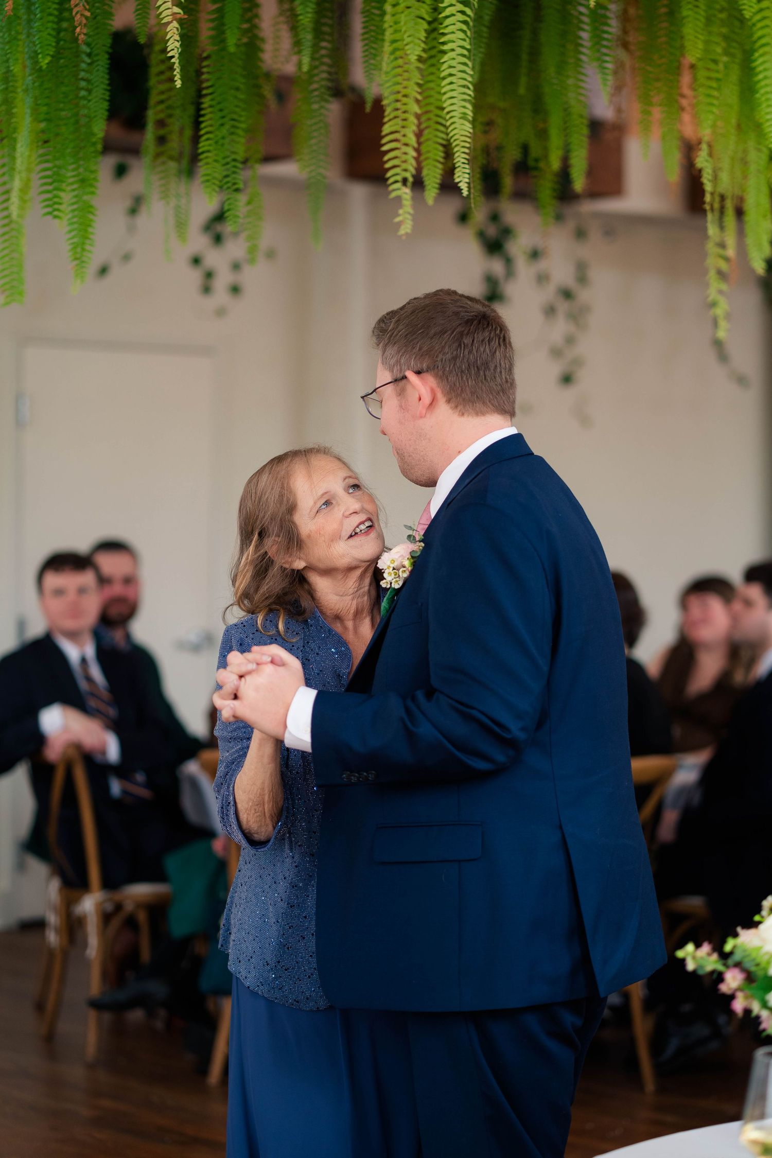 Groom in navy suit shares joyful mother-son dance at elegant wedding reception.