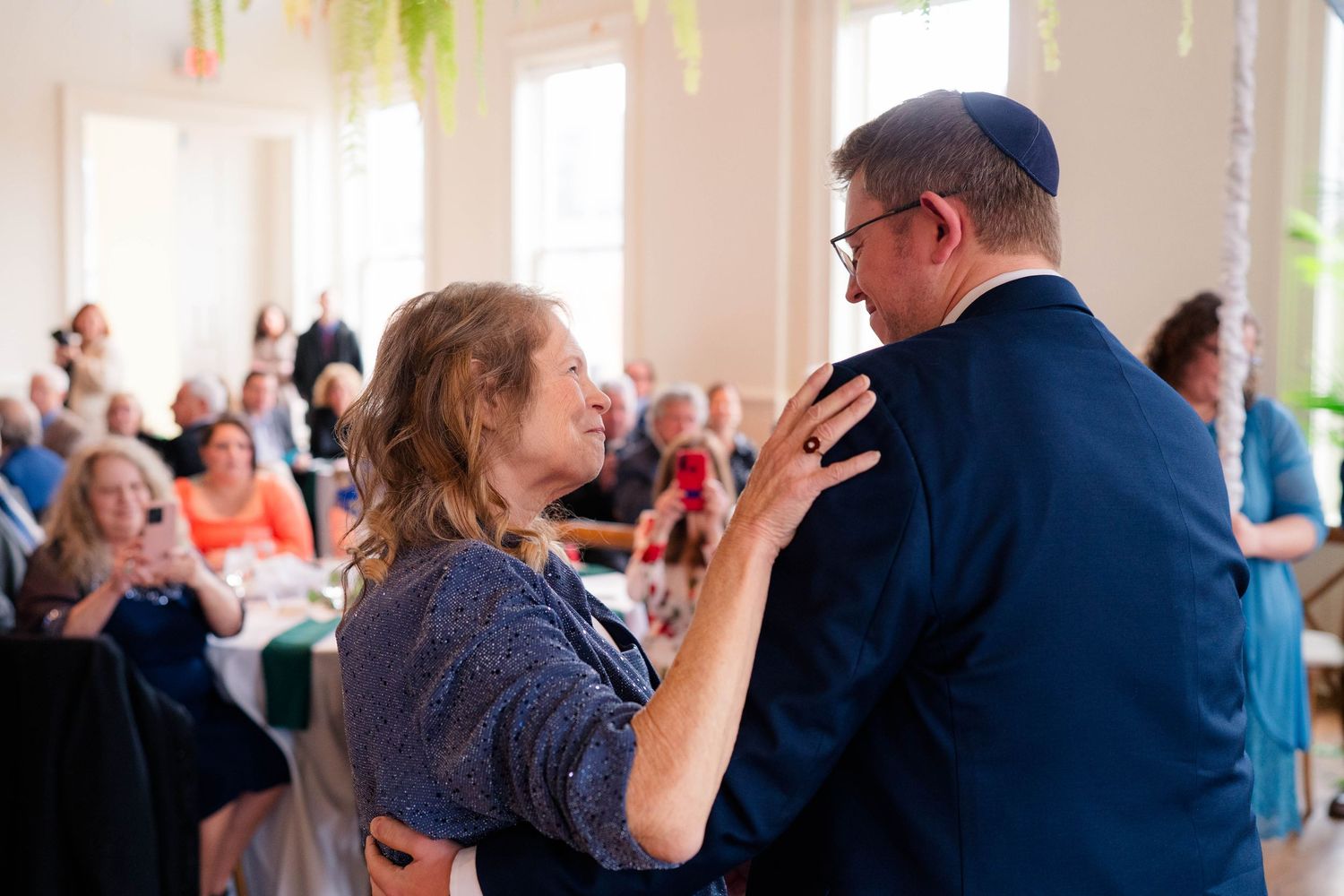 Mother and son share a tender dance at wedding reception with guests seated in background.