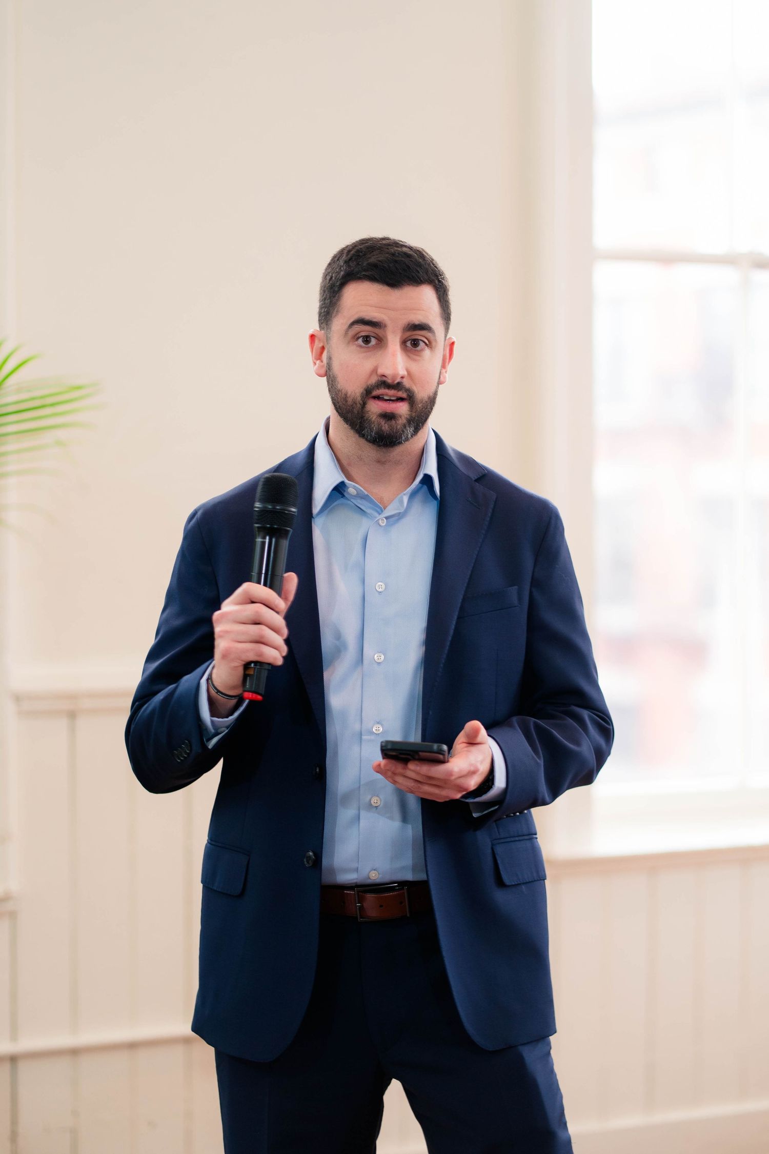 Bearded man in navy suit speaks into microphone while holding phone at wedding reception.