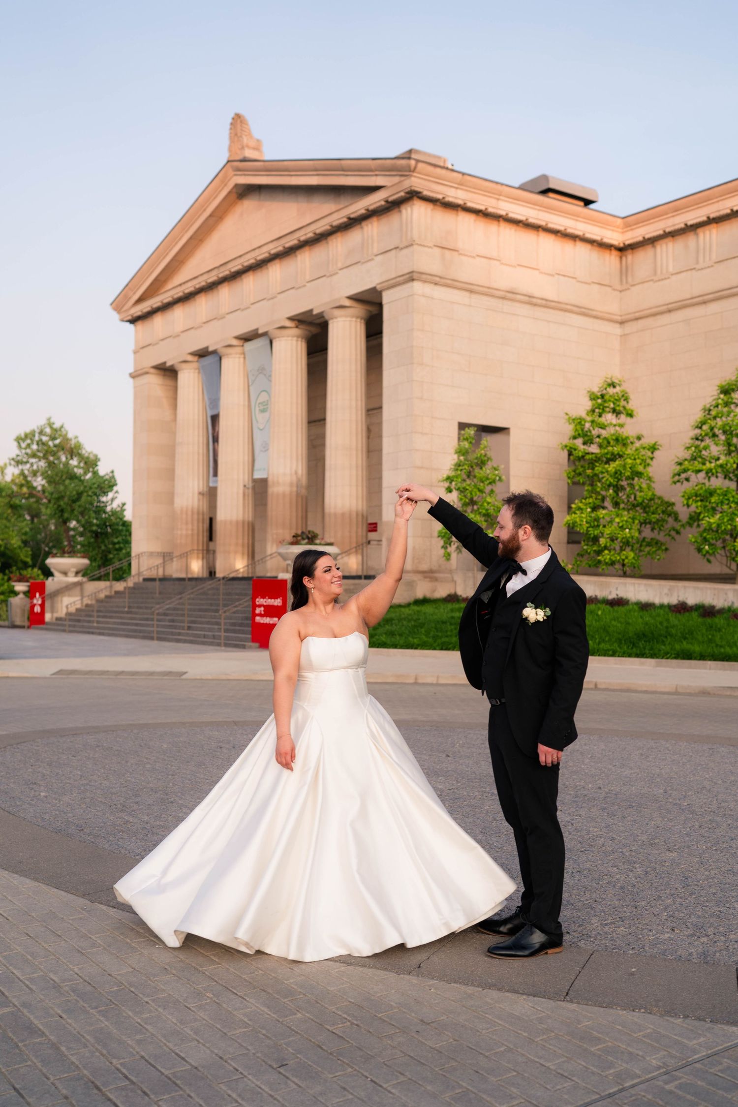 Groom twirls bride in a white ball gown in front of a neoclassical building at sunset.