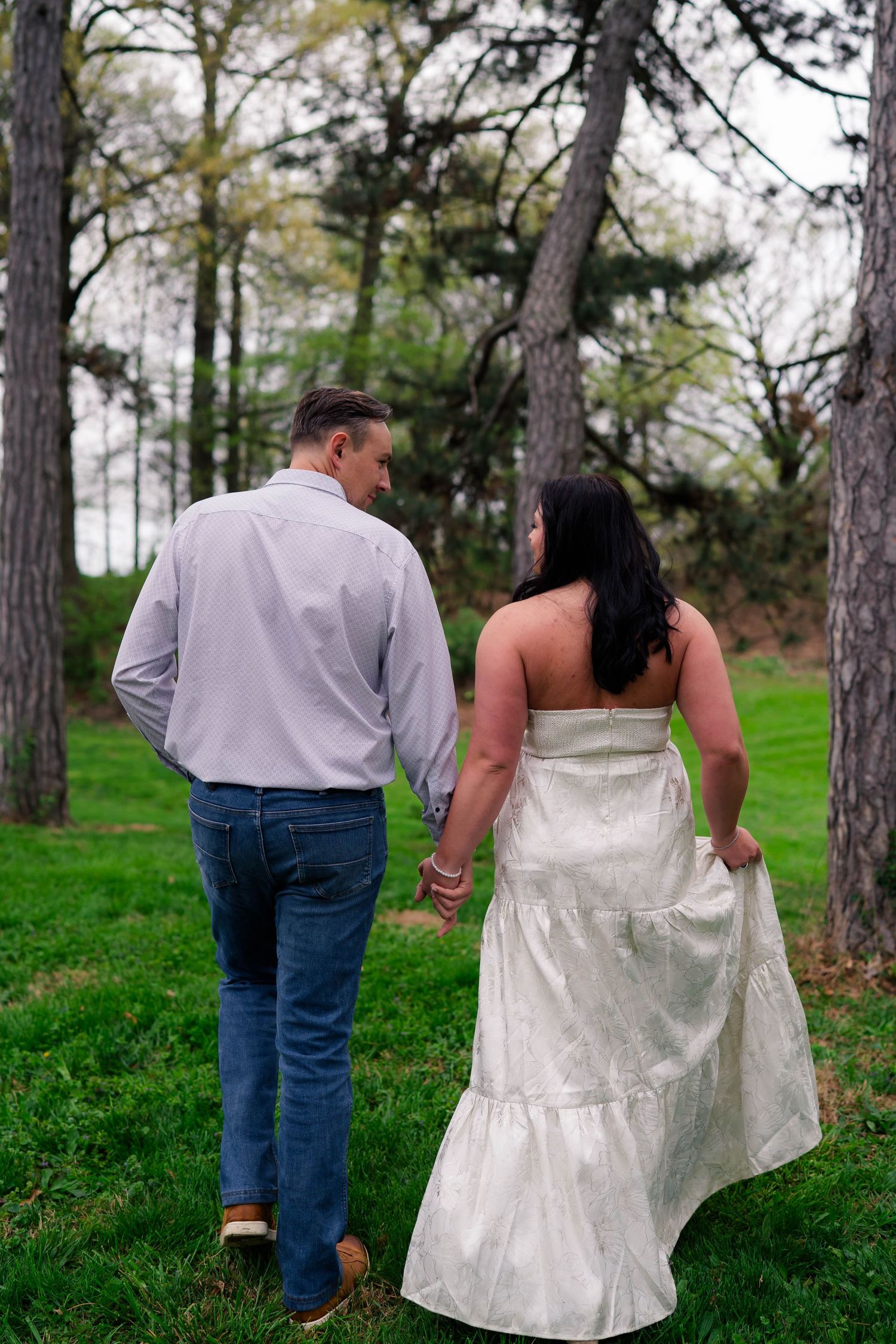Couple strolling hand in hand through green park, woman in white strapless dress glancing back at man in gray shirt.
