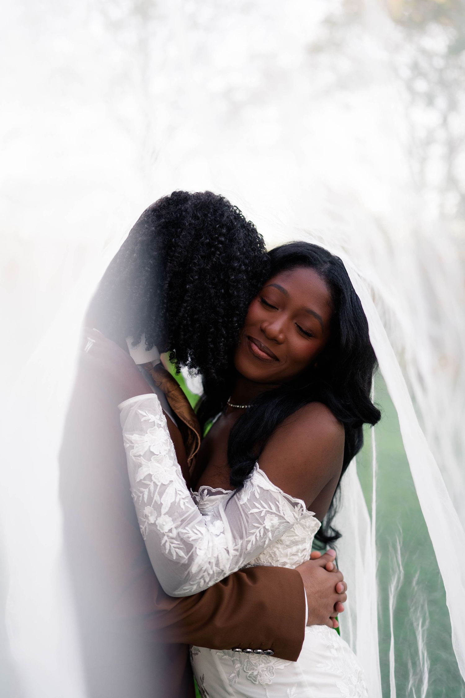 Two Black women in wedding attire share a tender embrace under a flowing white veil outdoors.