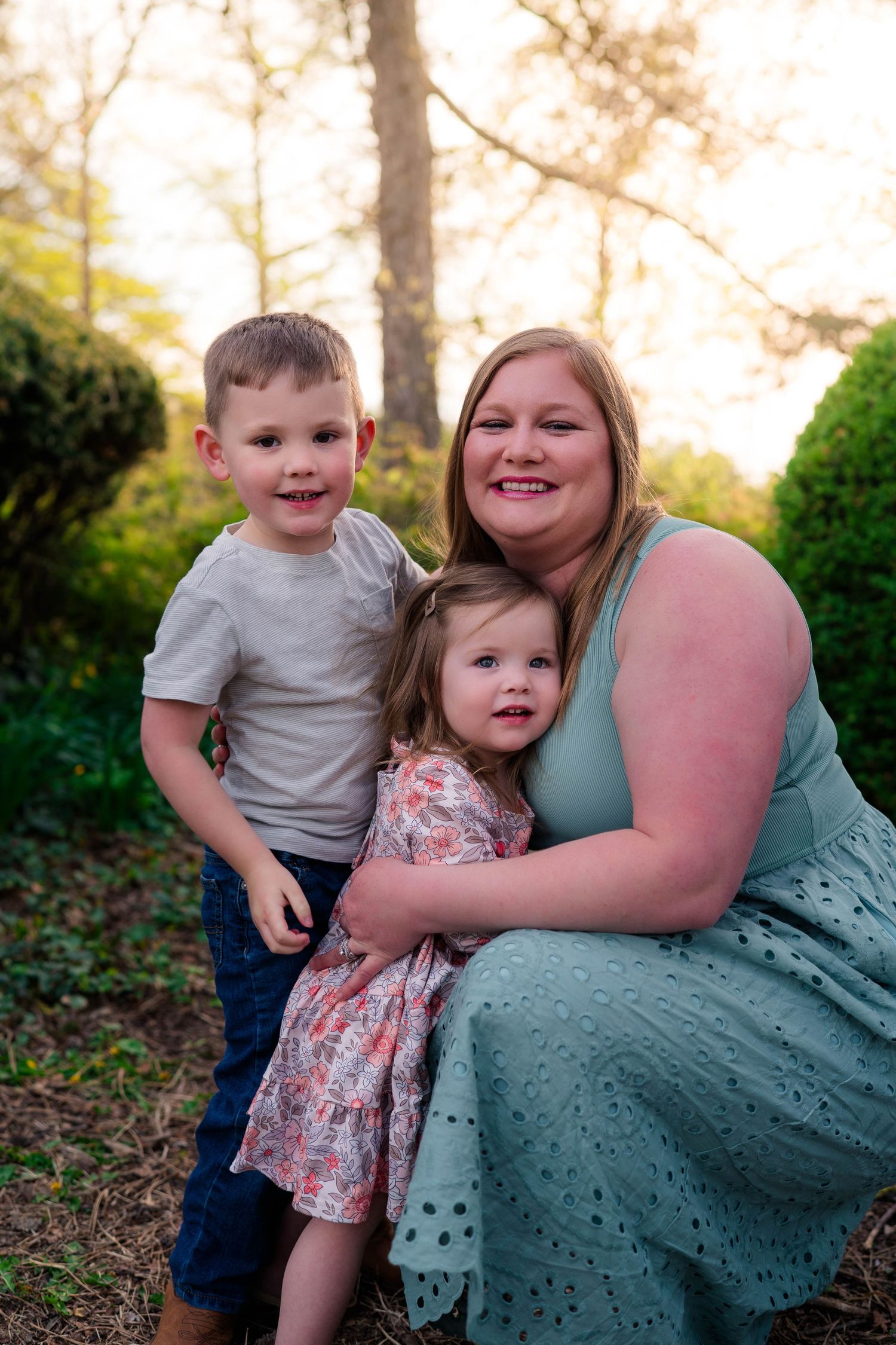 A smiling mother in a teal dress crouches outdoors with her young son and toddler daughter in a garden setting.