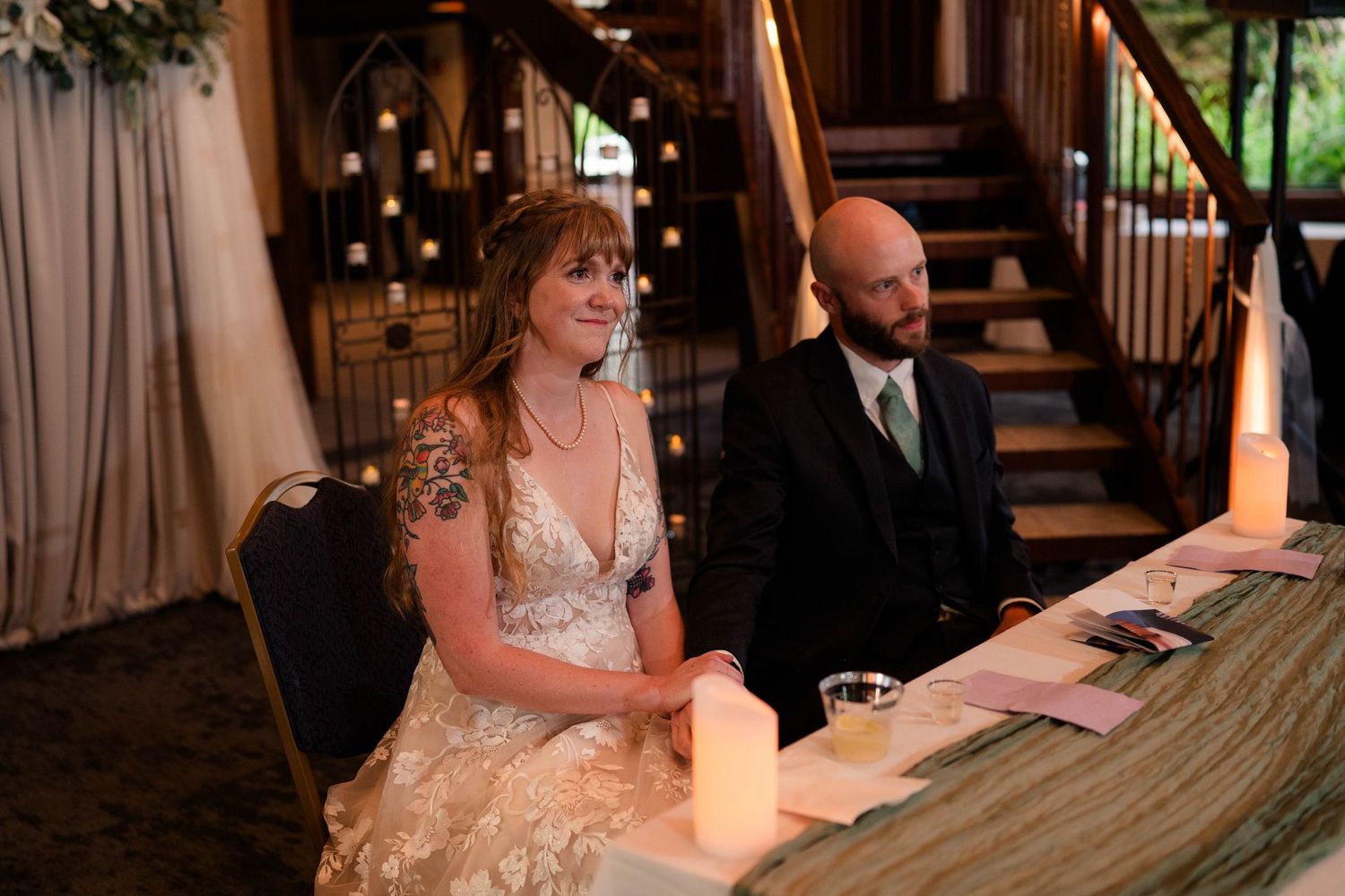 Bride and bald groom smile at sweetheart table with candles at wedding reception.
