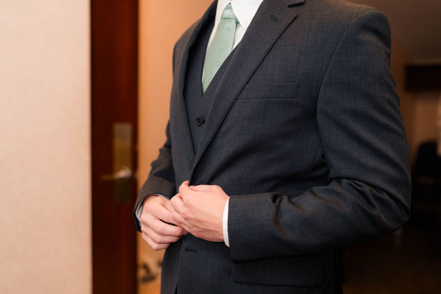 Man in dark three-piece suit adjusting jacket buttons, wearing light green tie and white dress shirt.