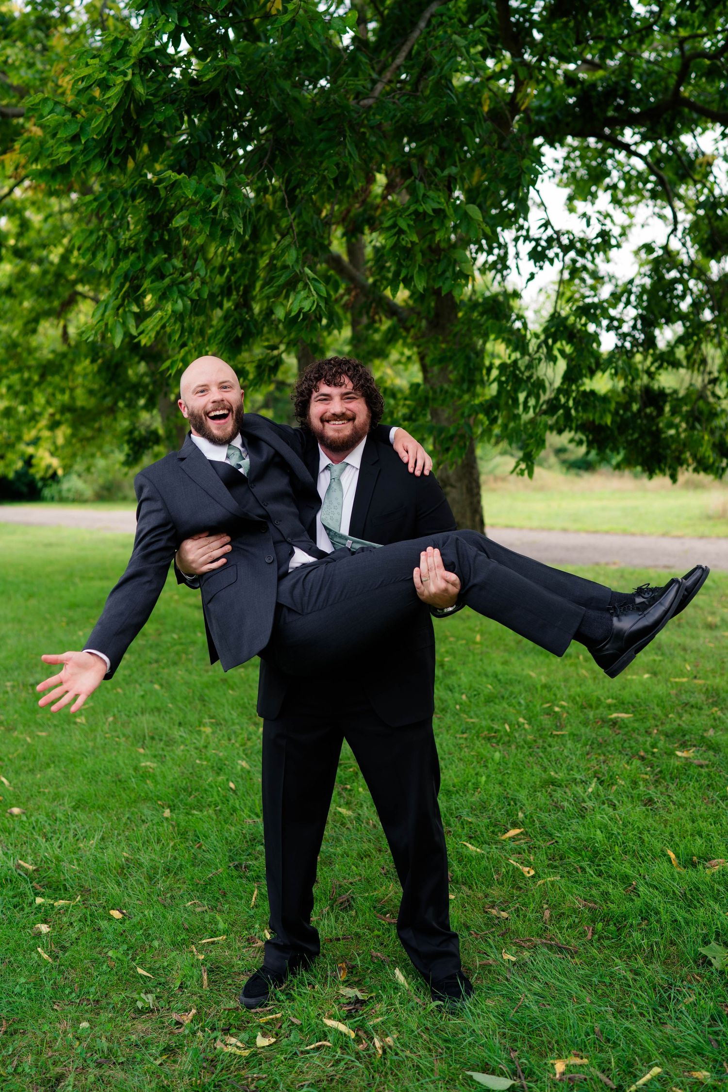 Two groomsmen laugh as one carries the other in his arms during fun outdoor wedding party photos in a park.